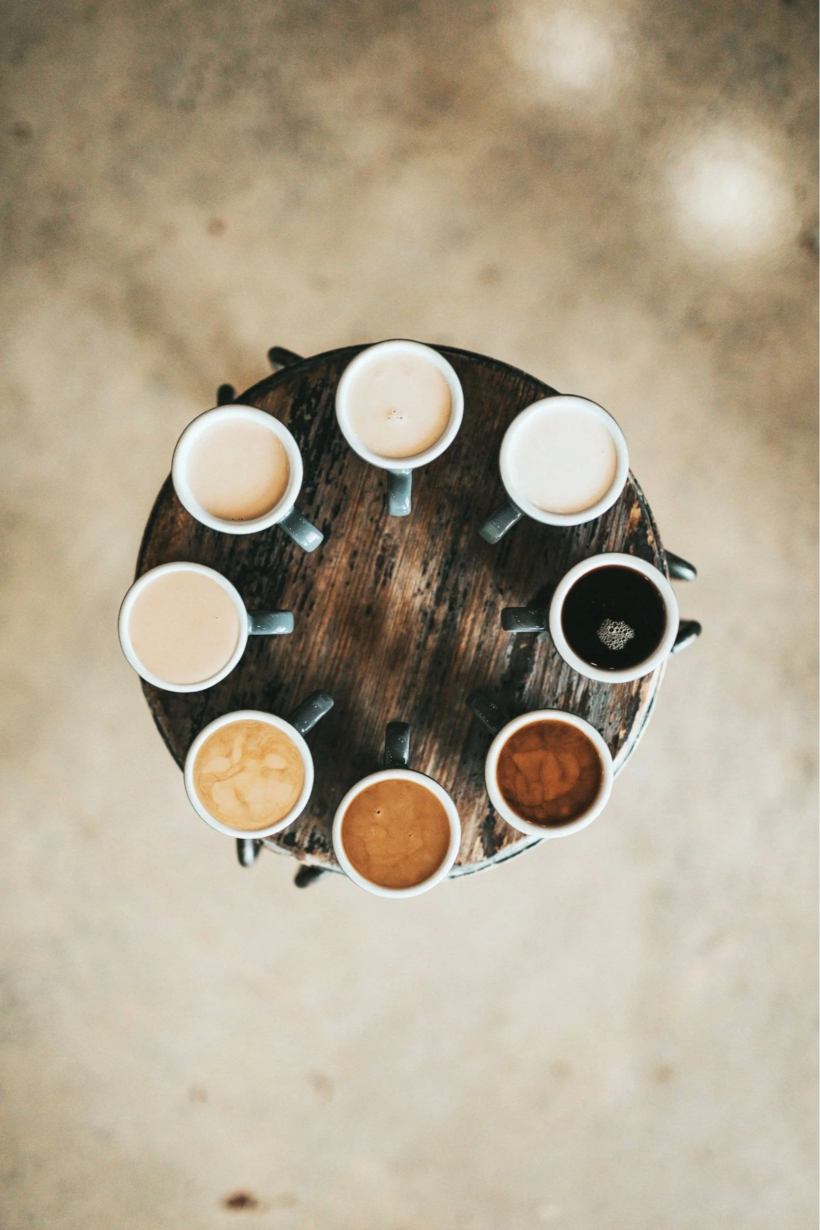Top-down view of a rustic wooden circular table with ten coffee cups filled with varying shades of coffee.