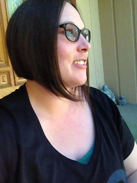 A woman with short black hair, wearing glasses, smiling, and looking to her right. She is dressed in a black top and is standing outside near a wooden door or wall.