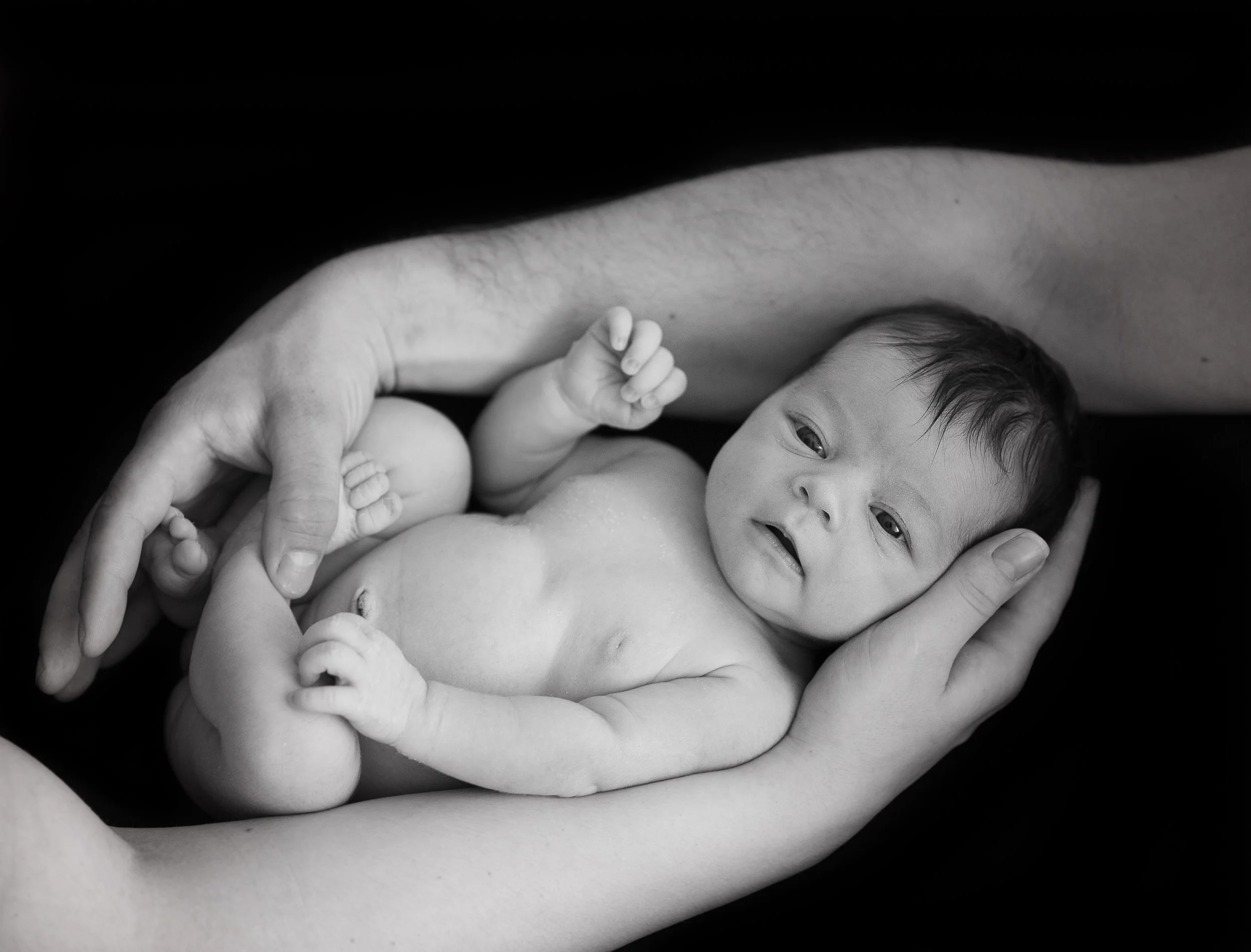 A baby lying on a bed, held gently by the hands of the mother and father, with a close-up of the baby's face and body, in black and white.