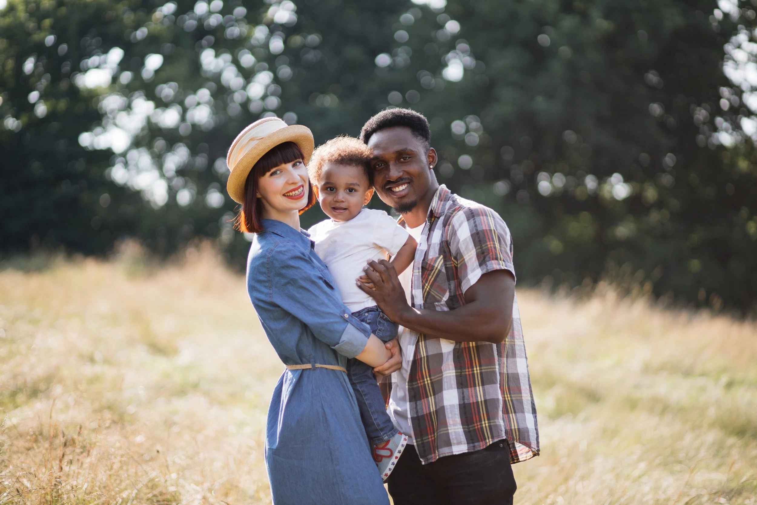 A happy family of four outdoors in a grassy area with trees in the background, including a woman with a hat, a man, a young girl, and a small boy.