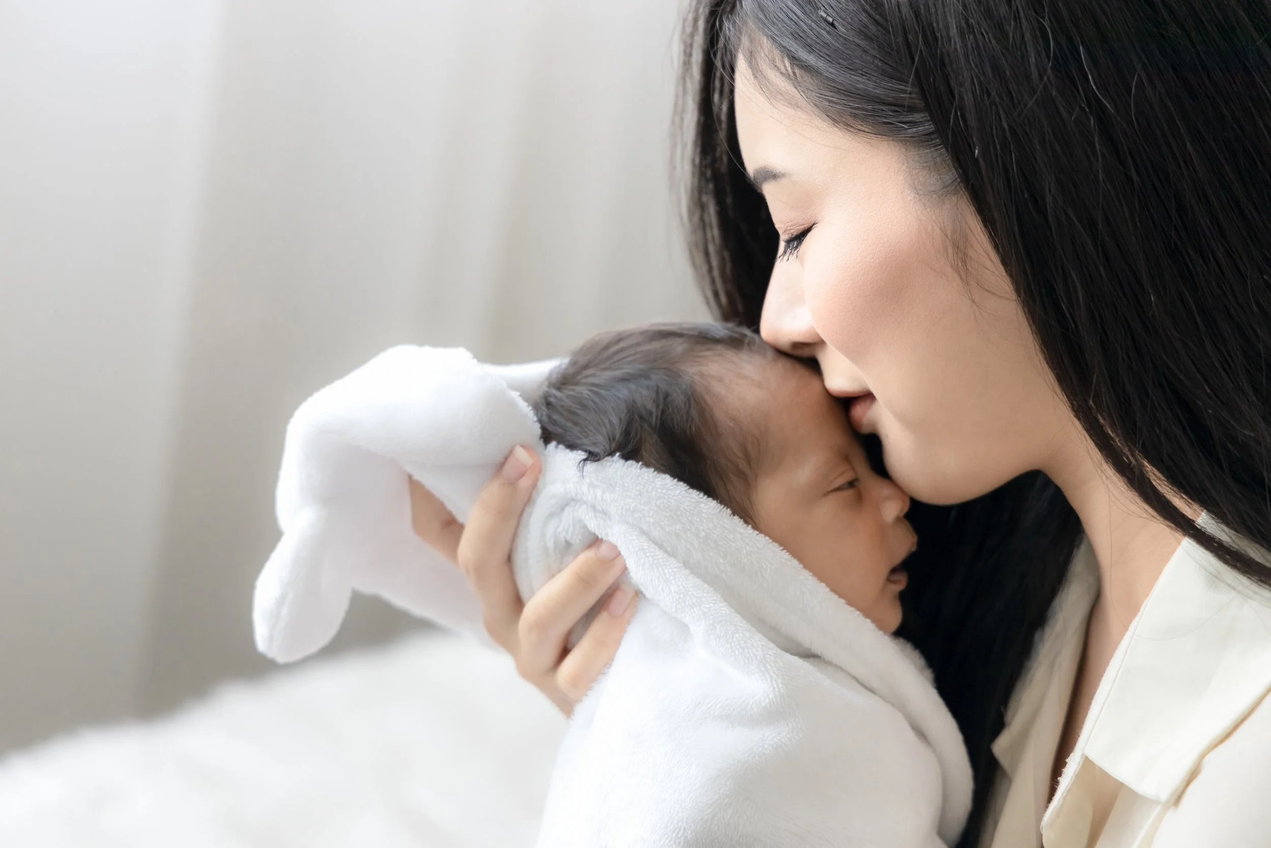 A woman with long dark hair gently holds and kisses a sleeping baby wrapped in a white blanket.