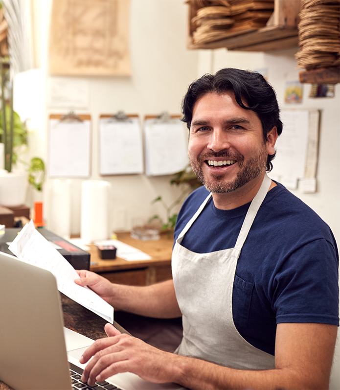 A smiling male small business owner with dark hair and a beard, wearing a navy blue shirt and white apron, working at a counter in a cozy cafe or bakery