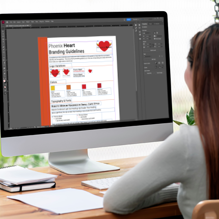 Woman working at a desk on a graphic design project about Phoenix Heart branding guidelines displayed on a computer screen, with a notebook and papers on the desk.