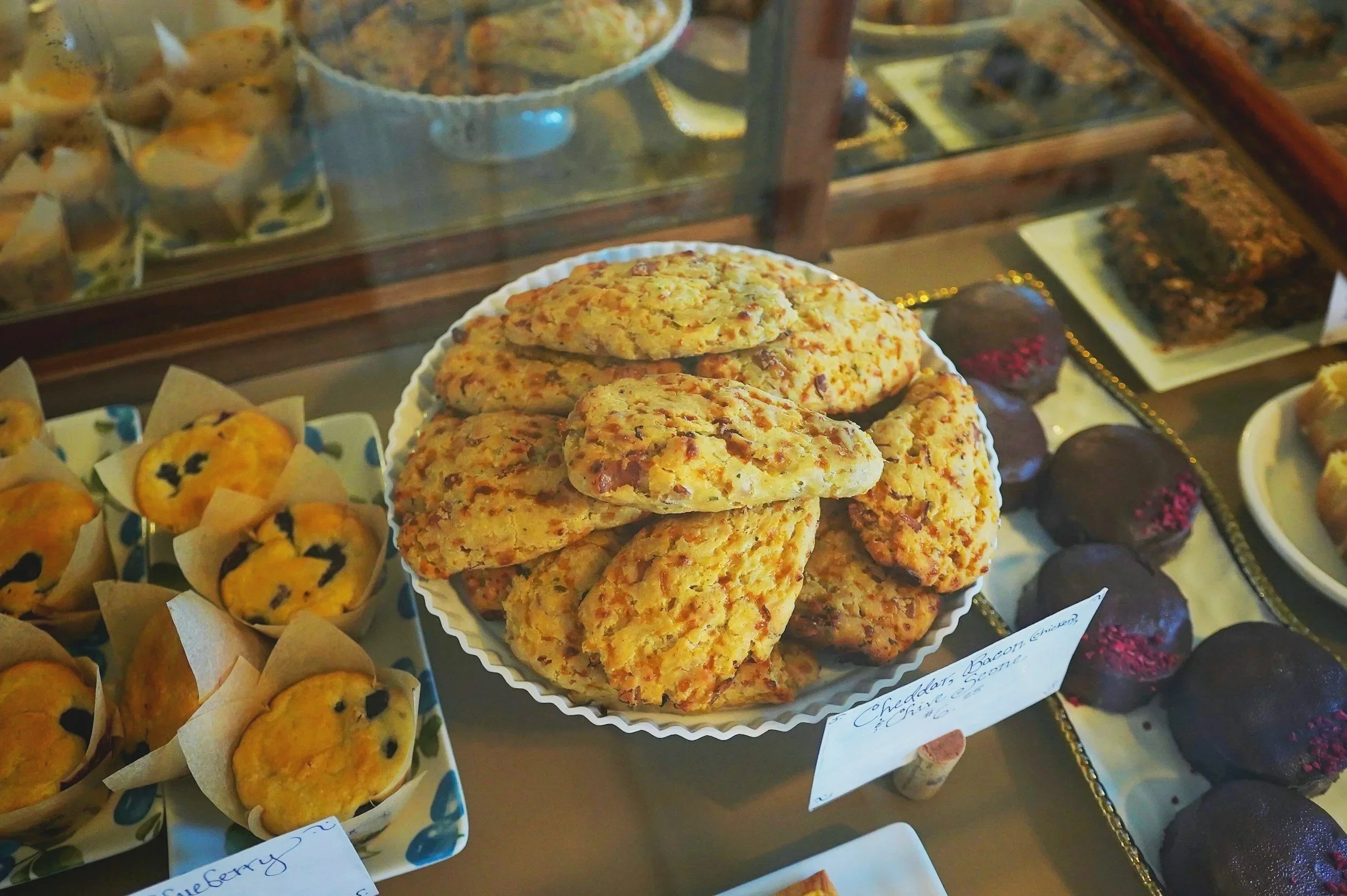 A display of various baked goods, including a stack of scones in the foreground, blueberry muffins on the left, and chocolate or berry cupcakes on the right, inside a glass display case.
