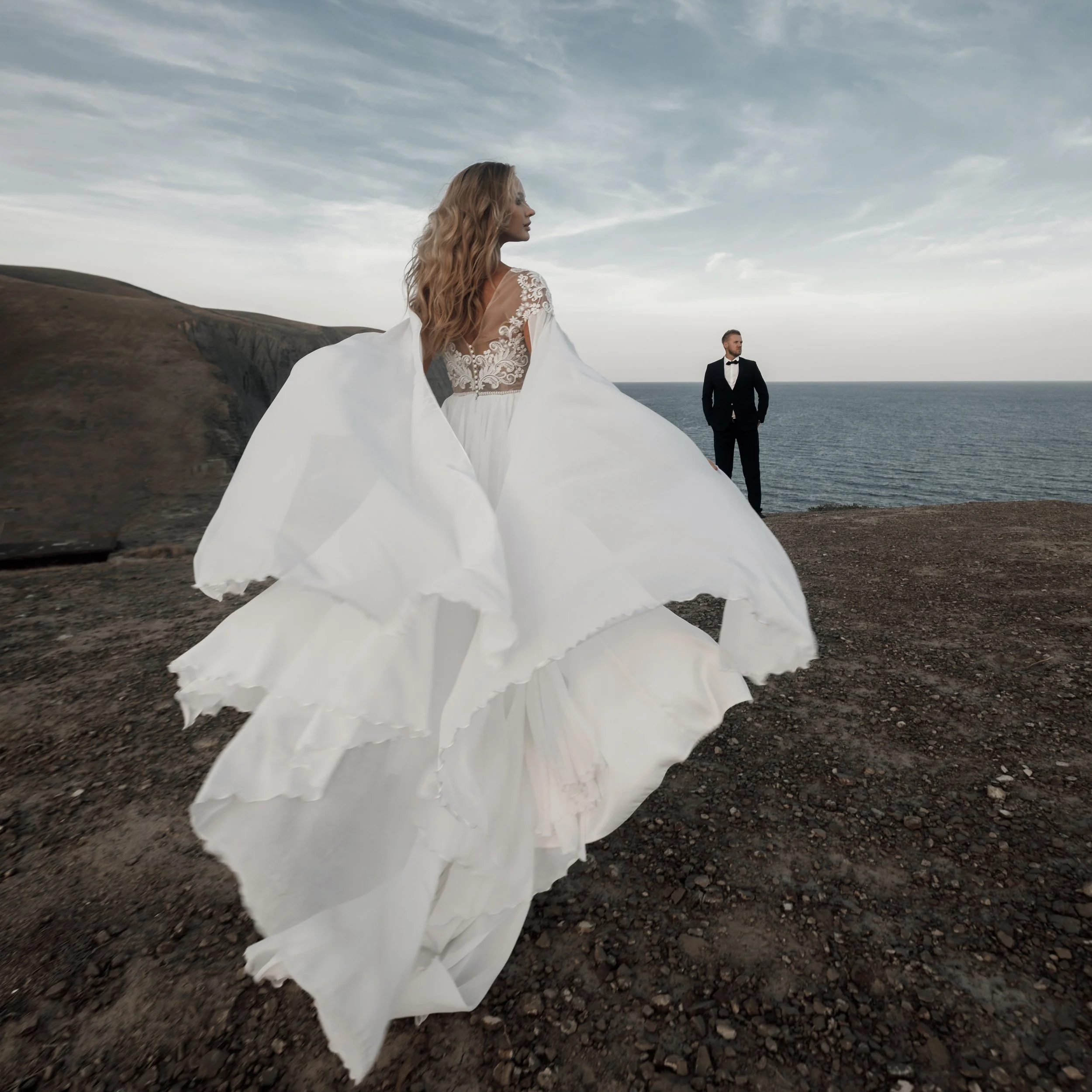 A woman in a white wedding dress with lace details stands on rocky terrain by the ocean, with a man in a black tuxedo in the background, under a cloudy sky.