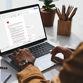 Person working on a laptop with a document open and being edited on the screen, surrounded by a potted plant and a wooden container with pencils, on a white desk.