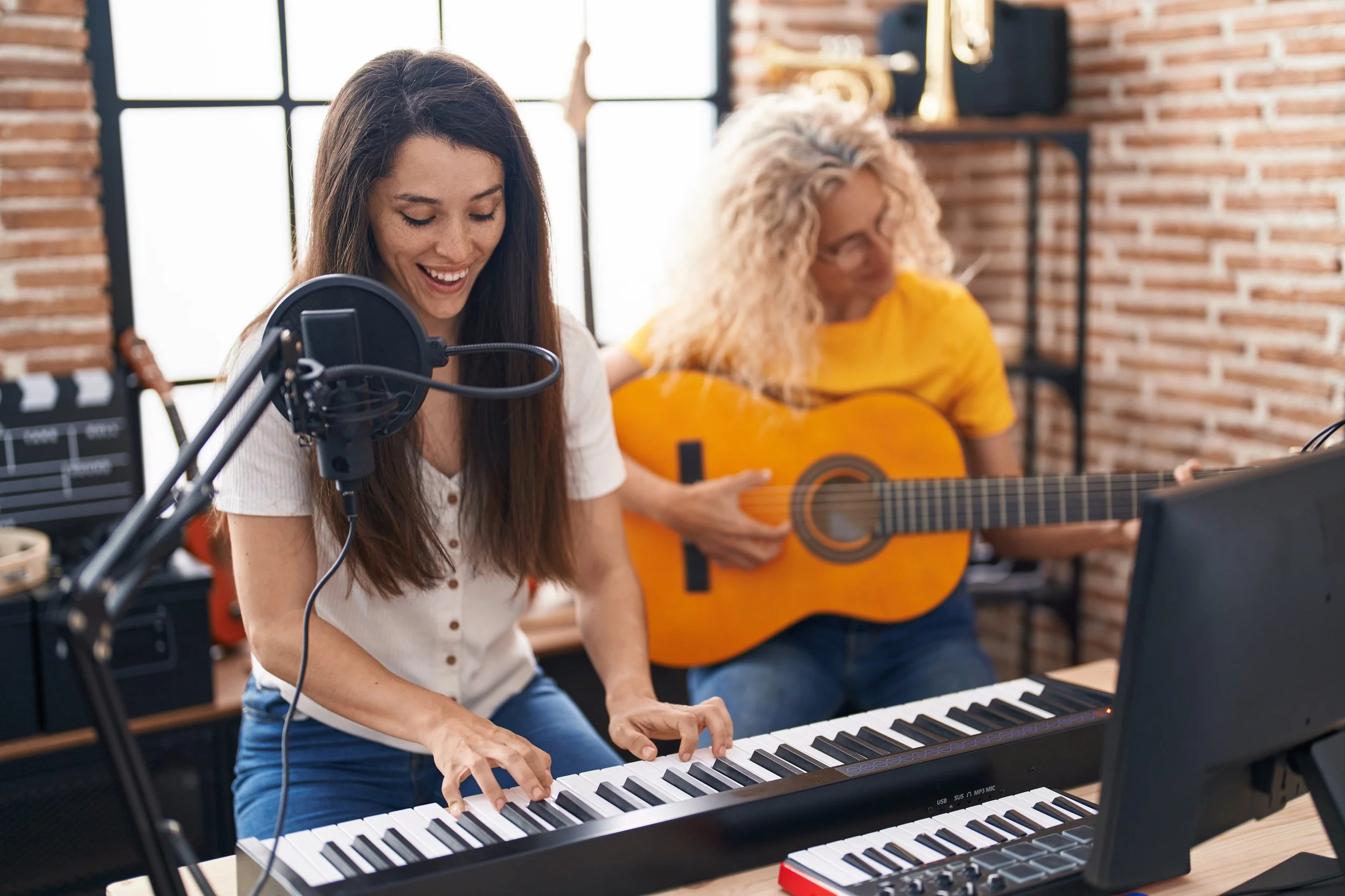 Two women playing musical instruments in a cafe with brick walls. One woman is playing the keyboard and the other is playing the guitar.