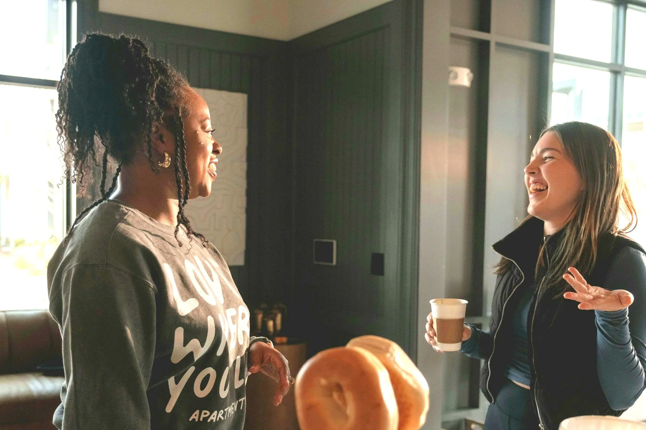 Two women are conversing indoors, one with dark skin, curly hair styled in a tall ponytail, wearing a gray sweatshirt, and the other with light skin, straight brown hair, wearing a black leather jacket, holding a coffee cup, and smiling.