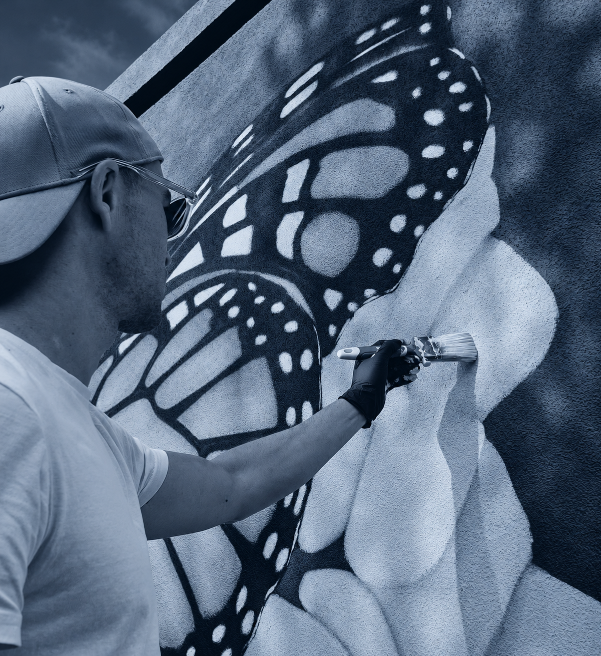 Un peintre de l'atelier Muréal réalise au pinceau les détails d'une fresque murale représentant un papillon monarque et des fleurs. Photographie aux tonalités bleu-gris.
