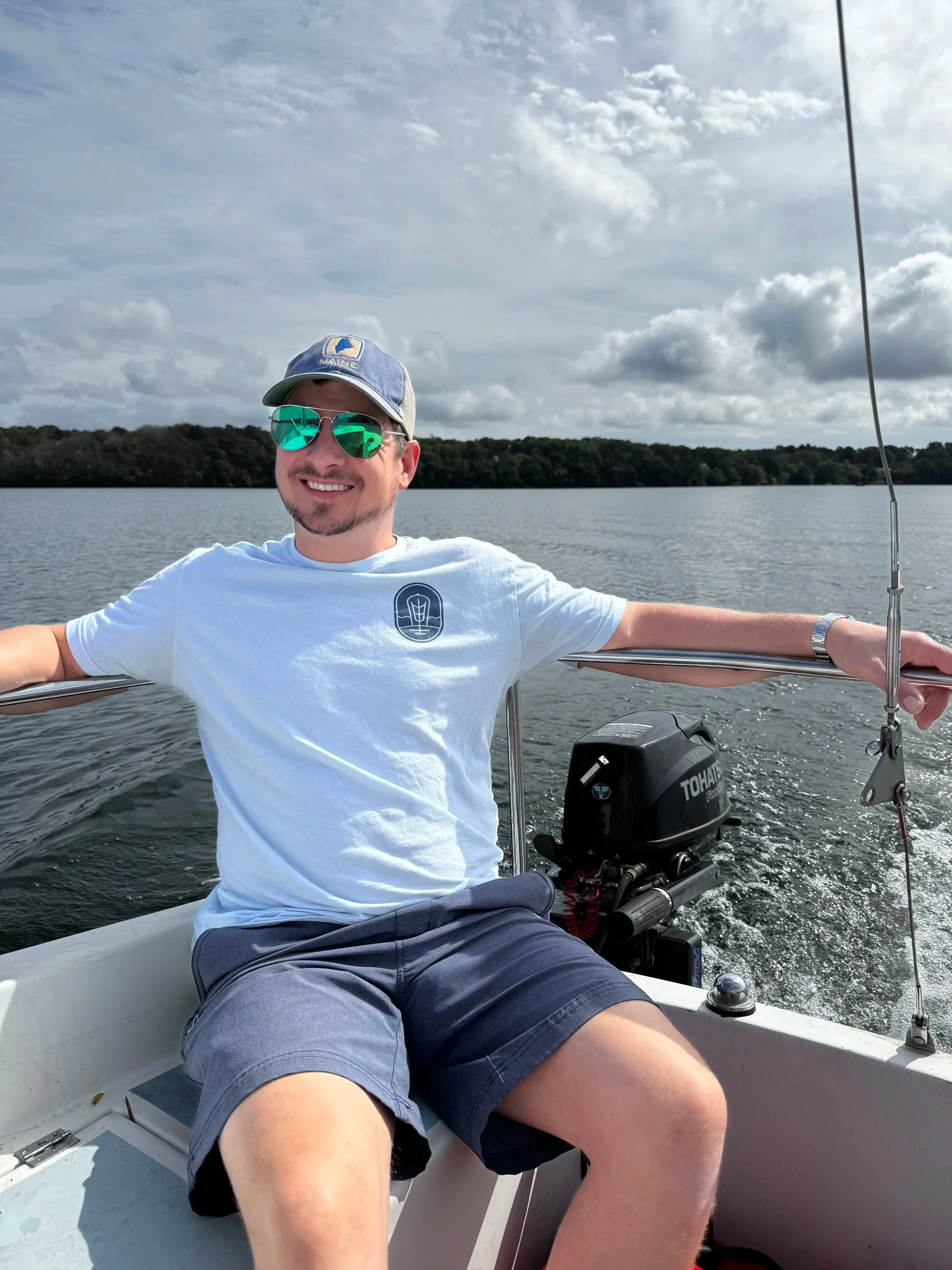 Smiling man wearing sunglasses and a baseball cap on a boat, with water and trees in the background.
