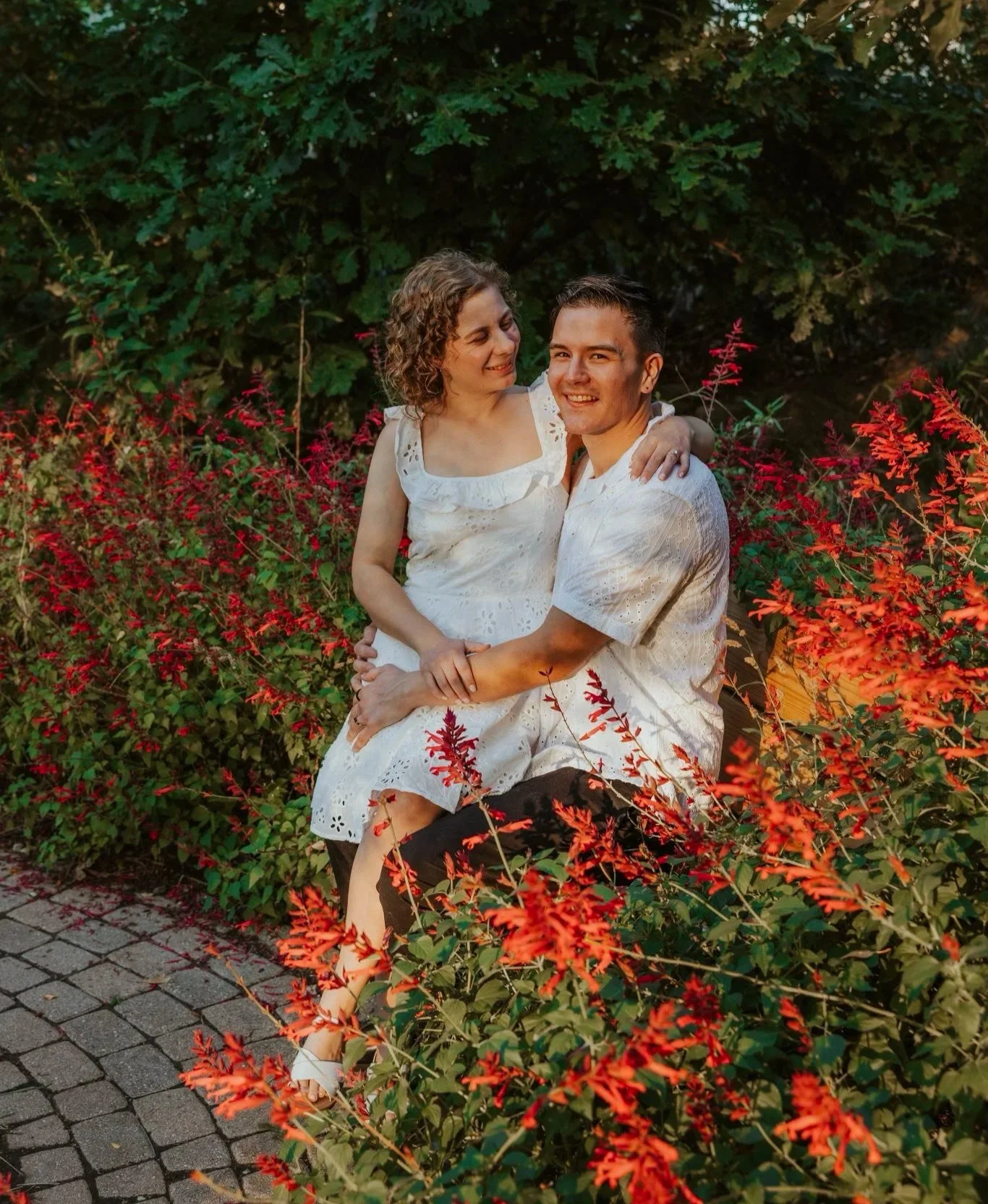 A couple sitting on a bench surrounded by red and green foliage, smiling and looking at each other. They are dressed in white summer outfits, with the woman resting her arm on the man's shoulder.