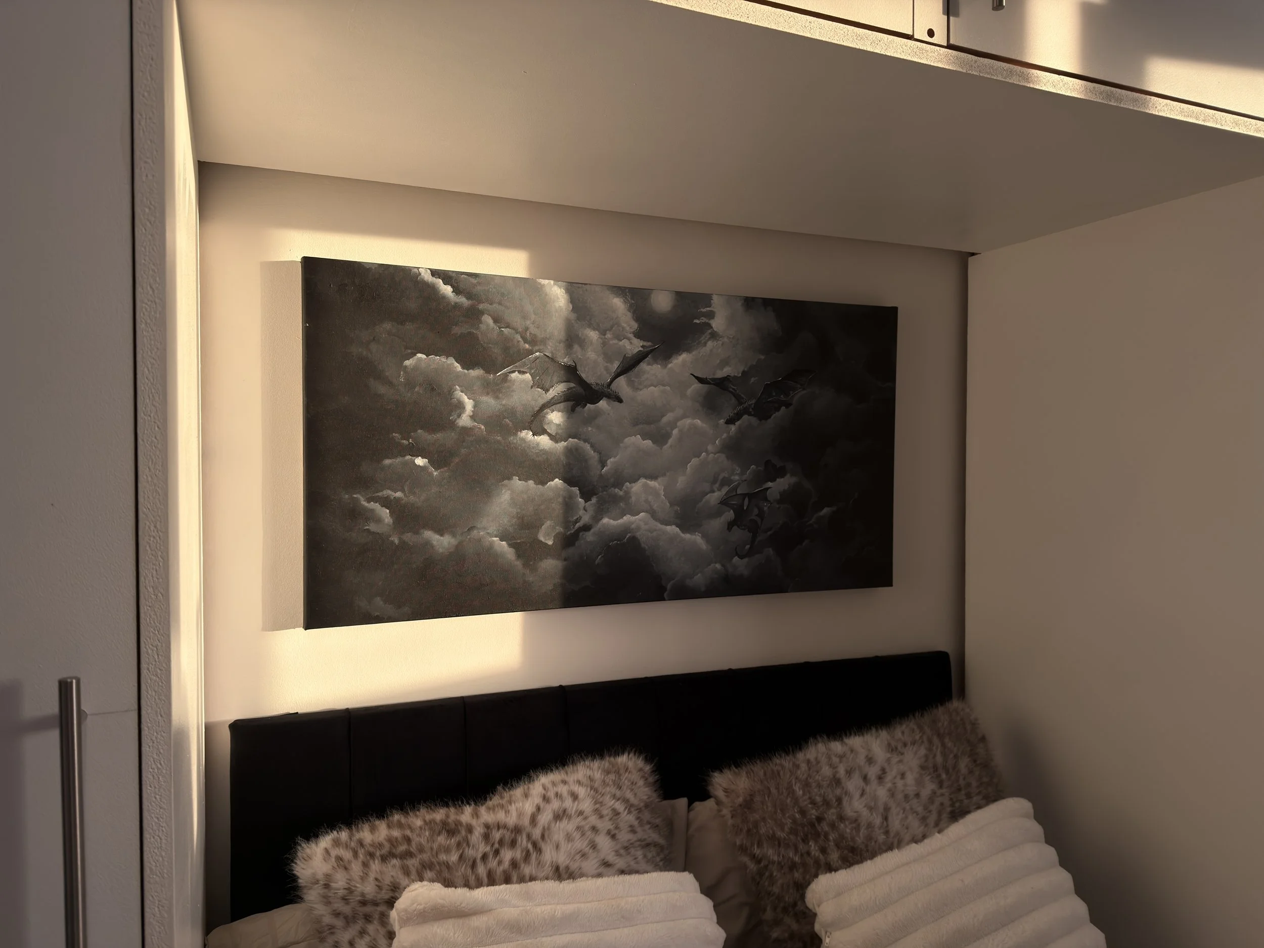 Black and white fantasy painting print by Alessia called 'Moonlit Drift' showing three dragons flying through the clouds with a hushed, mystical world below and moonlight illuminating a path through the sky. Featured in a bedroom with the sunset.