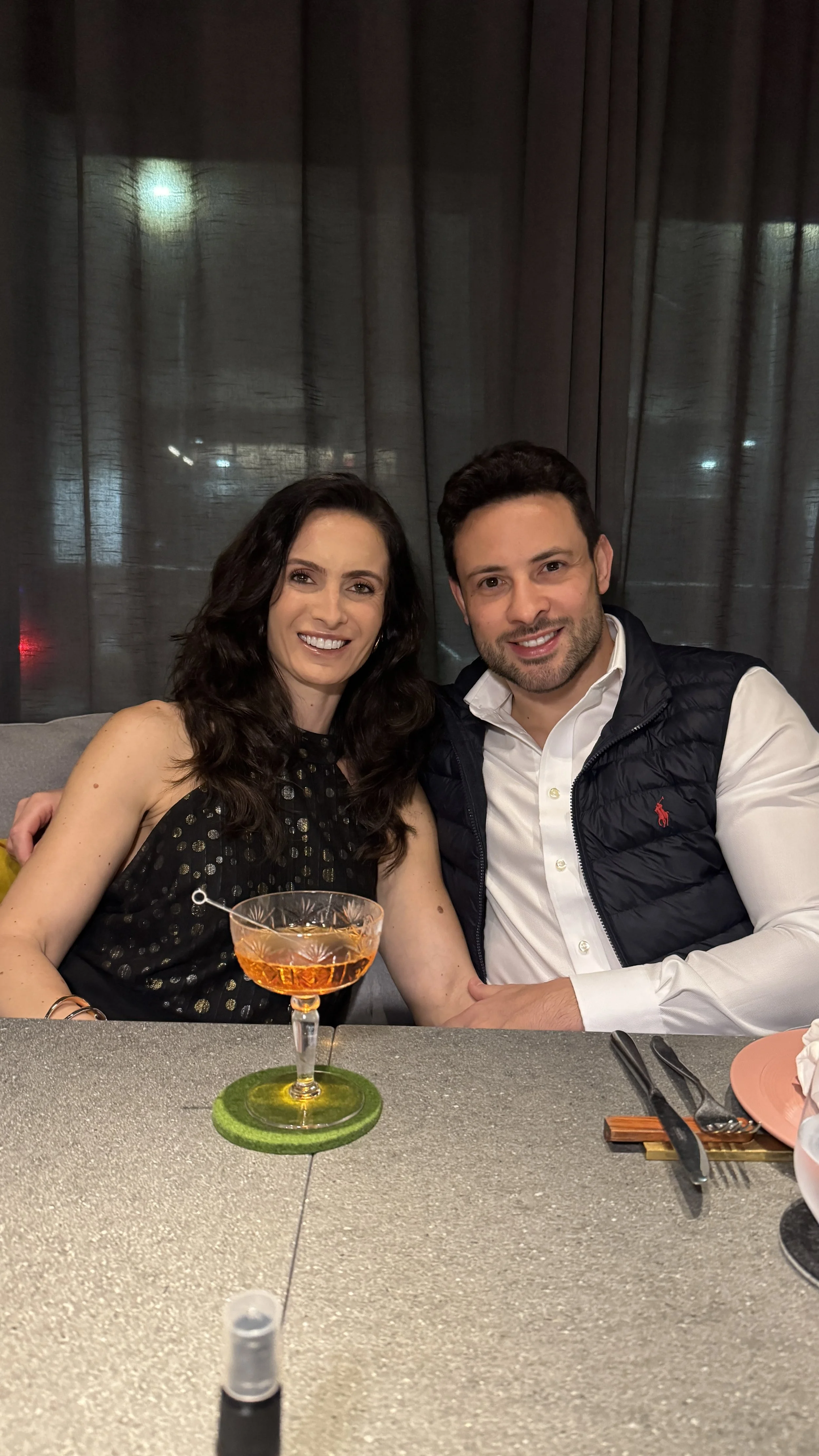 Casal sorridente sentado à mesa em um restaurante, com uma bebida colorida servida em uma taça na frente, fundo com cortinas escuras.