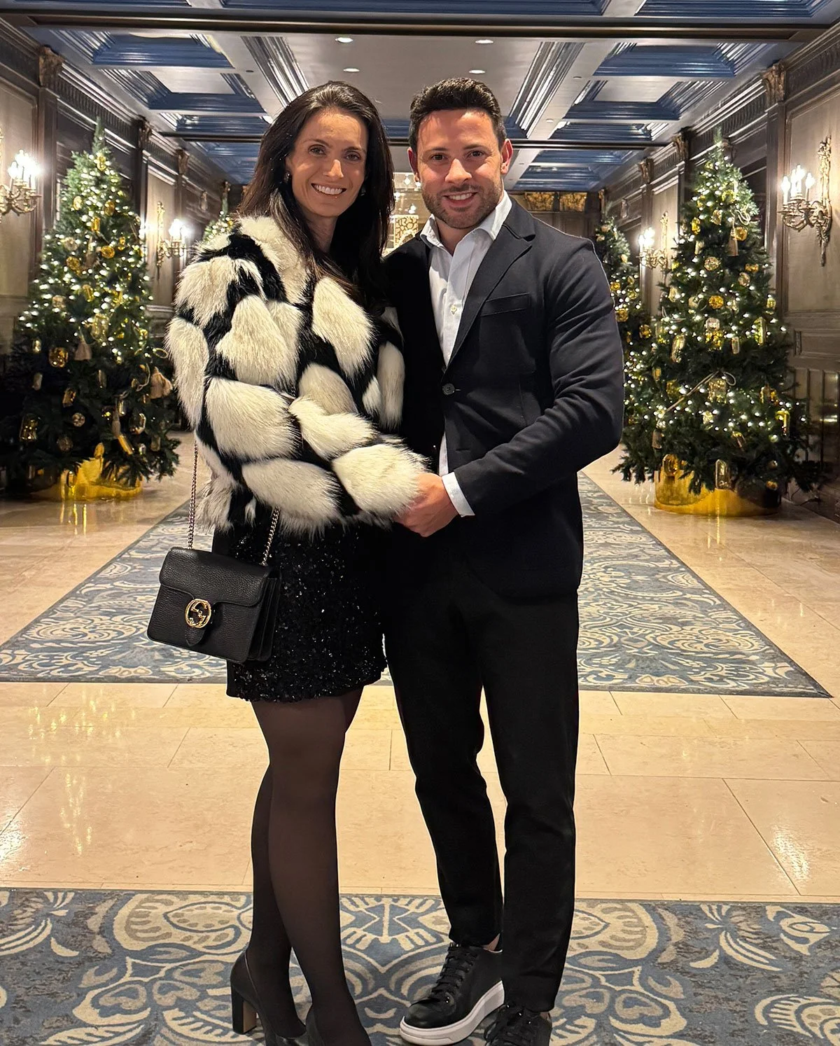 Um casal sorridente, vestindo roupas elegantes, segurando as mãos um do outro, em um ambiente decorado com árvores de Natal e luzes, possivelmente numa festa de fim de ano.