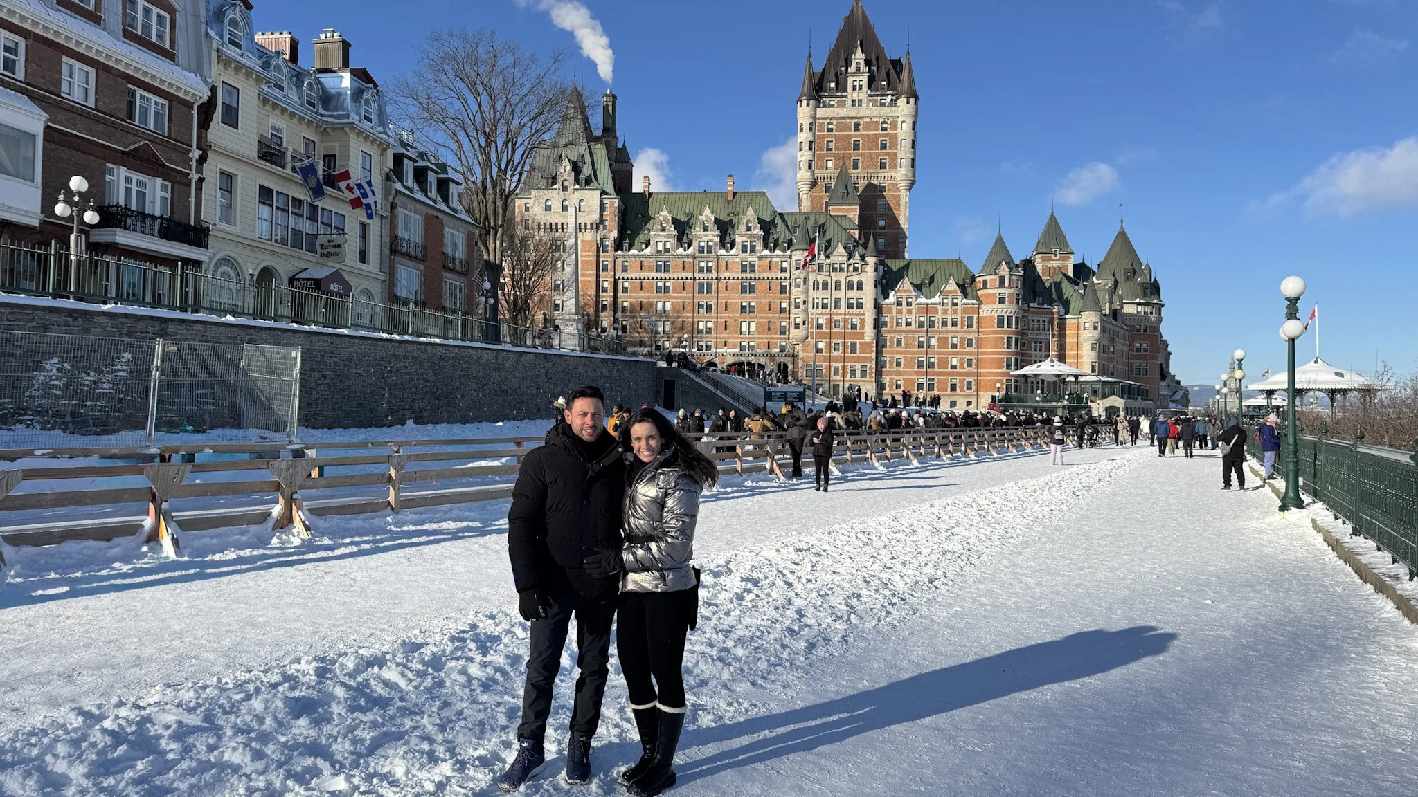 Casal feliz posando na neve com castelo ao fundo, céu claro e outros visitantes caminhando na praça.