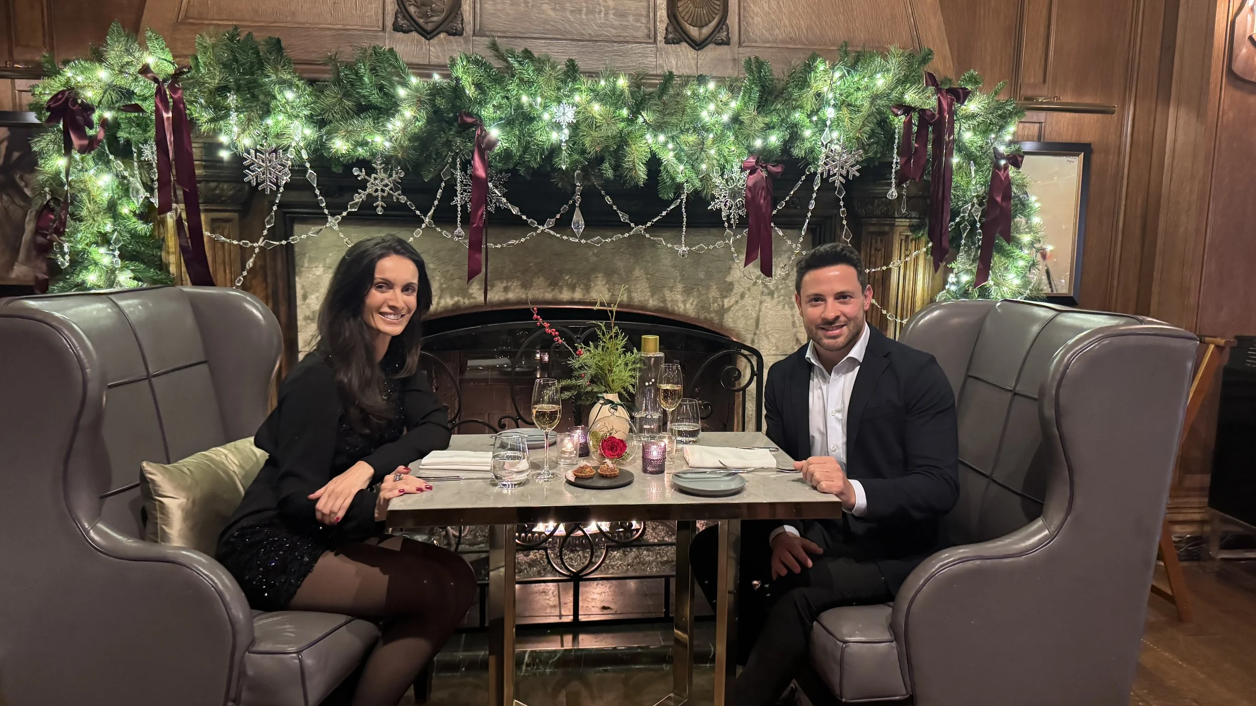 Homens e mulheres sentados em um restaurante decorado para o Natal, com uma lareira ao fundo decorada com guirlandas, luzes, fitas de cetim e snowflakes de vidro, compartilhando uma refeição.