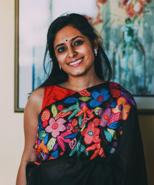 Smiling woman wearing a colorful embroidered dress with floral patterns, standing in front of an abstract painting.