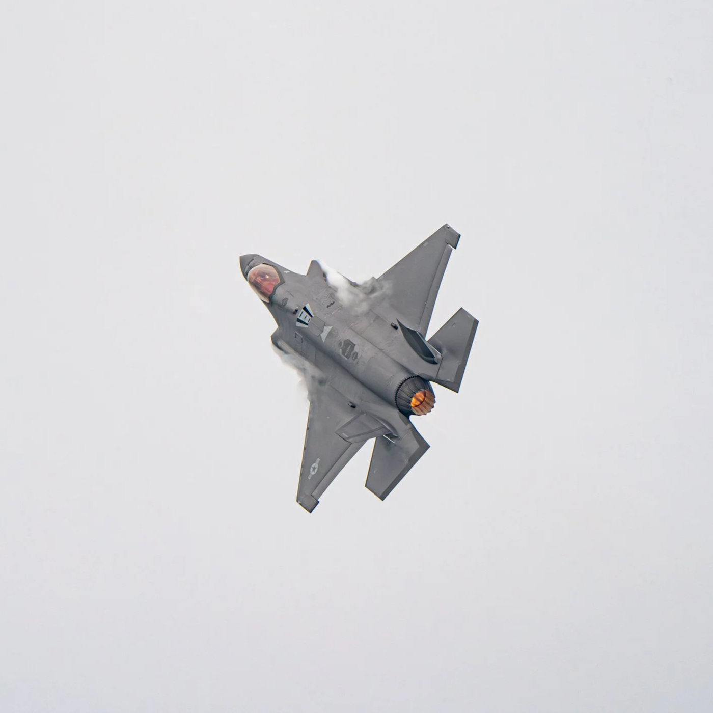 Ein militärisches Kampfjägerflugzeug fliegt durch den Himmel, Rauch und Flammen aus dem Triebwerk. Das Flugzeug ist grau und hat eine fortschrittliche, aerodynamische Form.