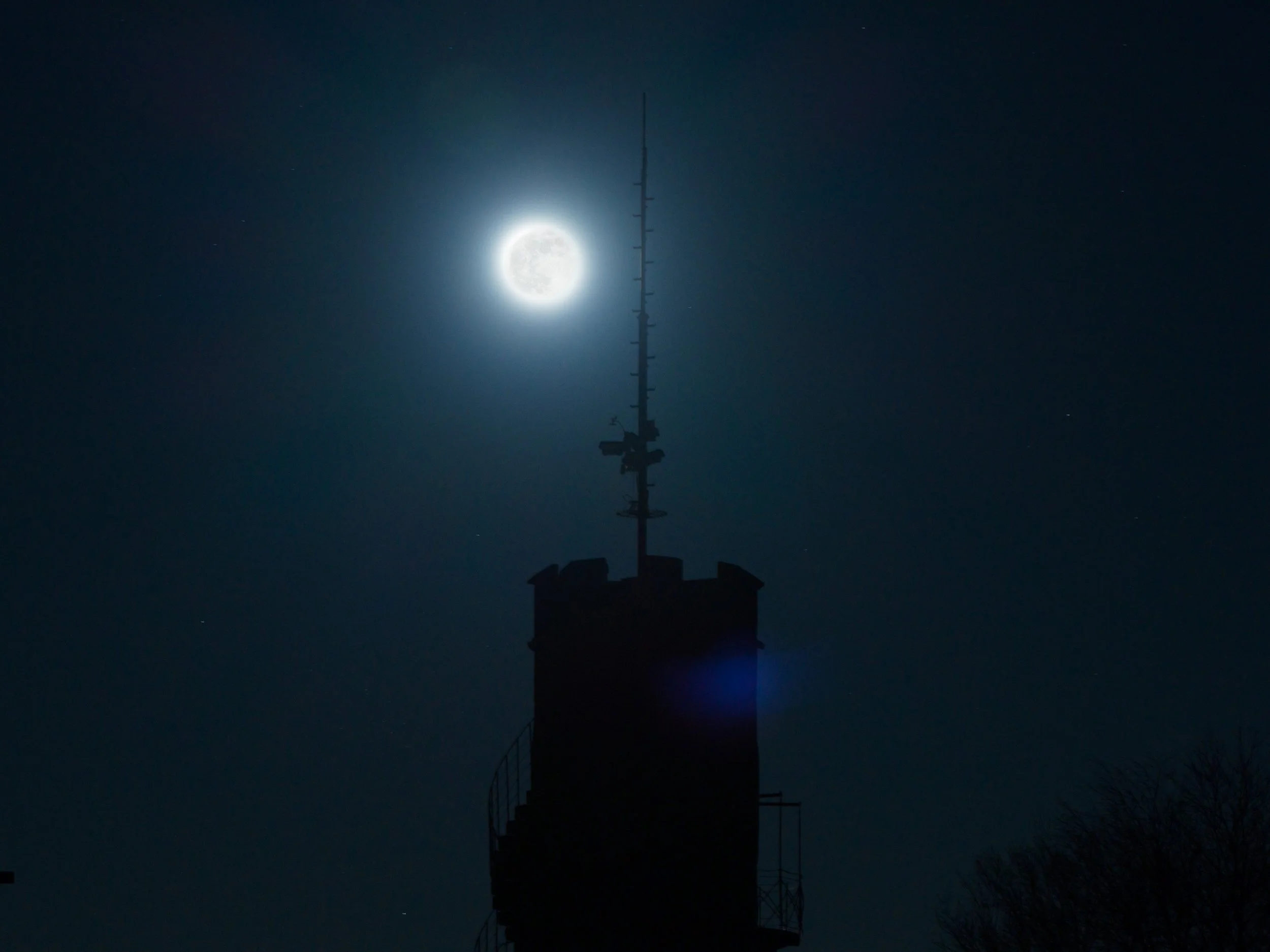 Vollmond hinter einem Sendeturm bei Nacht, mit dunklem Himmel und wenigen Sternen.