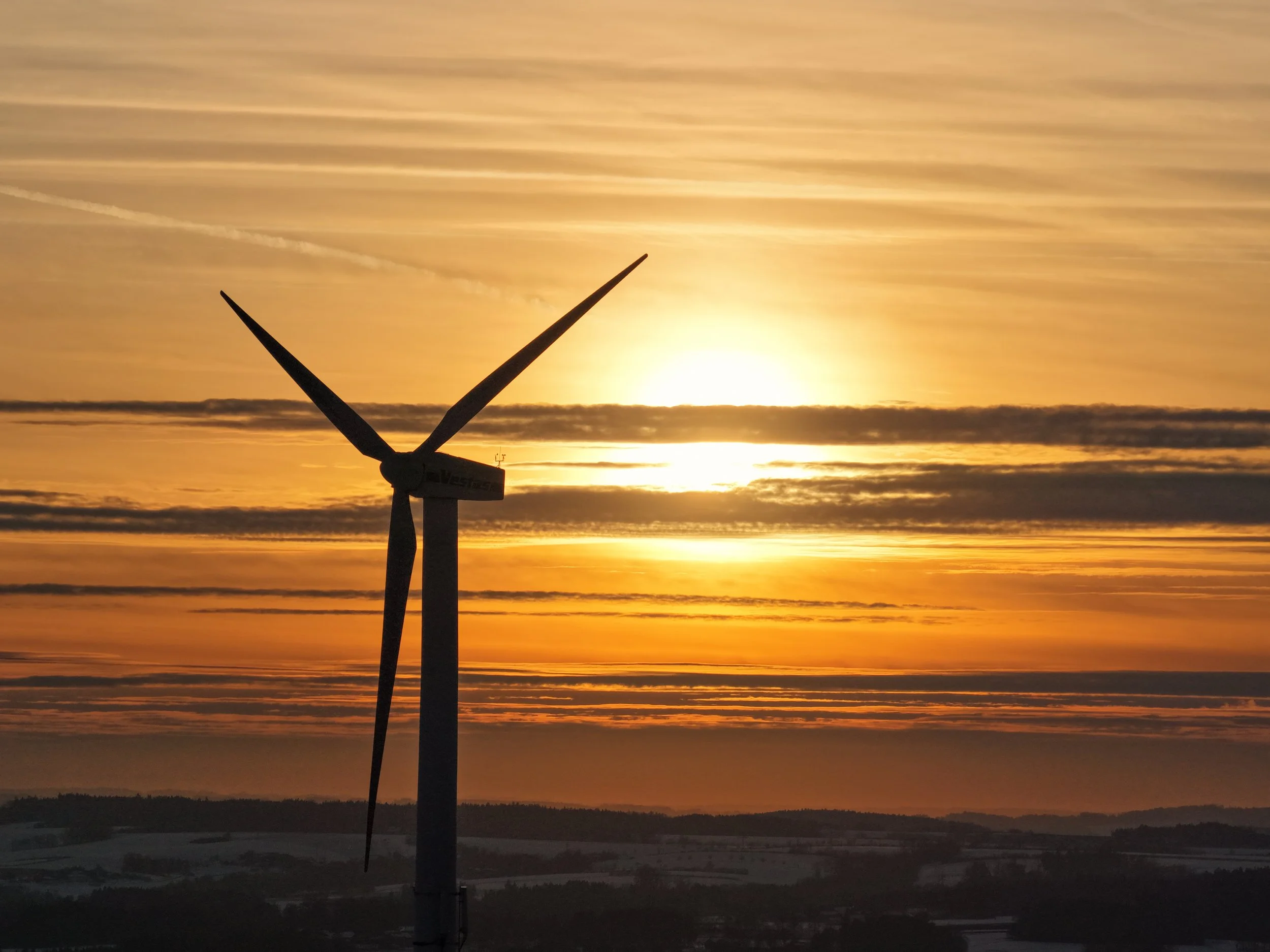 Ein Windrad vor einem Sonnenuntergang mit bewölktem Himmel und einer Landschaft im Hintergrund.