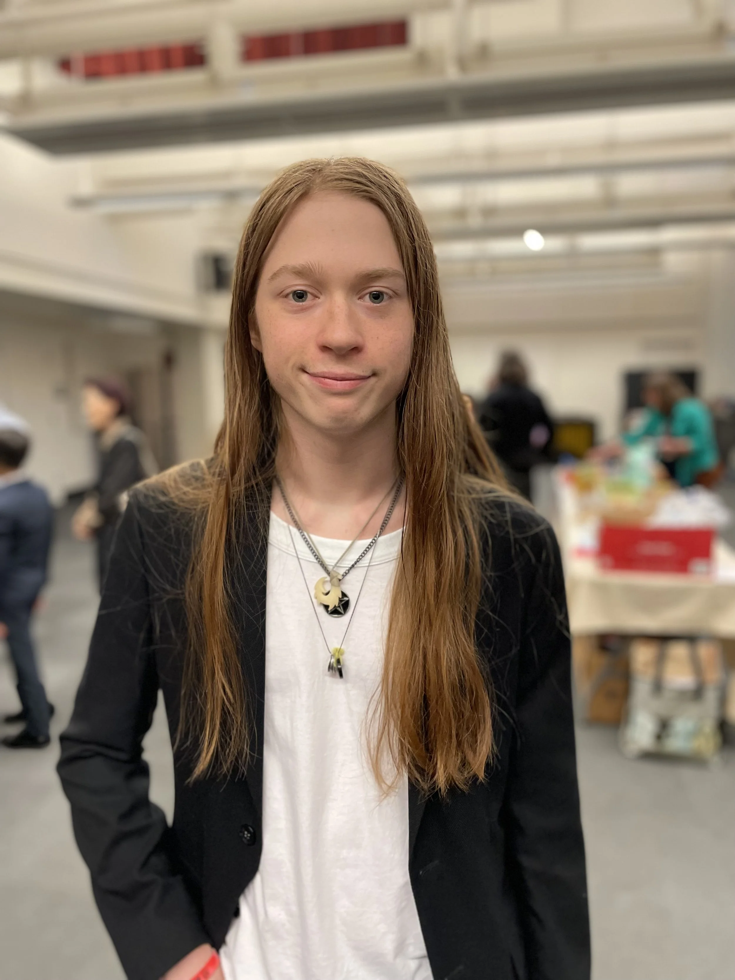 A young person with long red hair, wearing a black blazer over a white shirt, standing in a busy indoor setting with people and tables in the background.