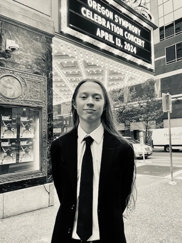 A young person with long hair wearing a suit and tie standing outside next to a marquee sign that reads "Oregon Symphony Celebration Concert April 13, 2024" in an urban area.