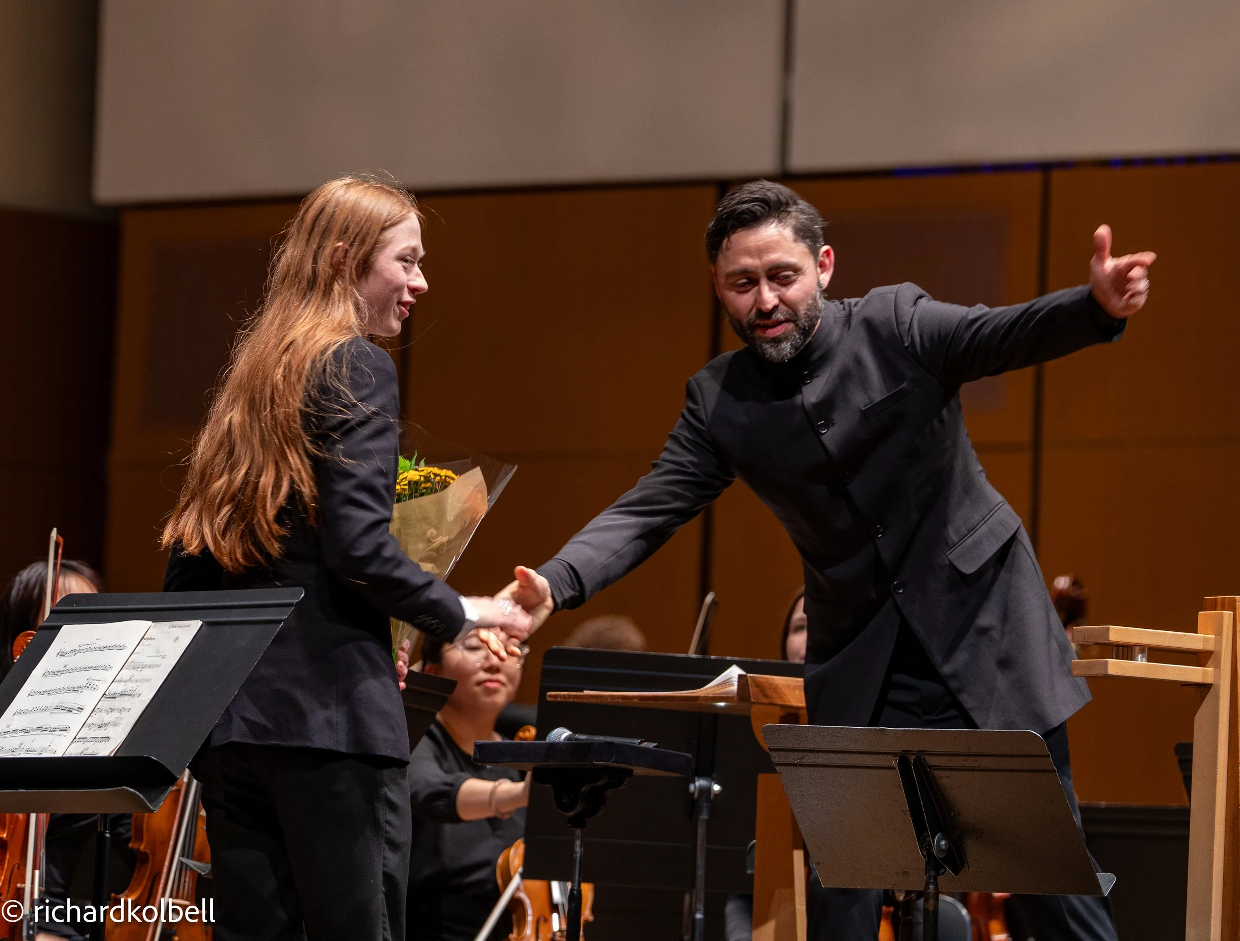 A man is shaking hands with a woman holding a bouquet of flowers on stage during a formal concert or award ceremony, with other musicians seated behind them.