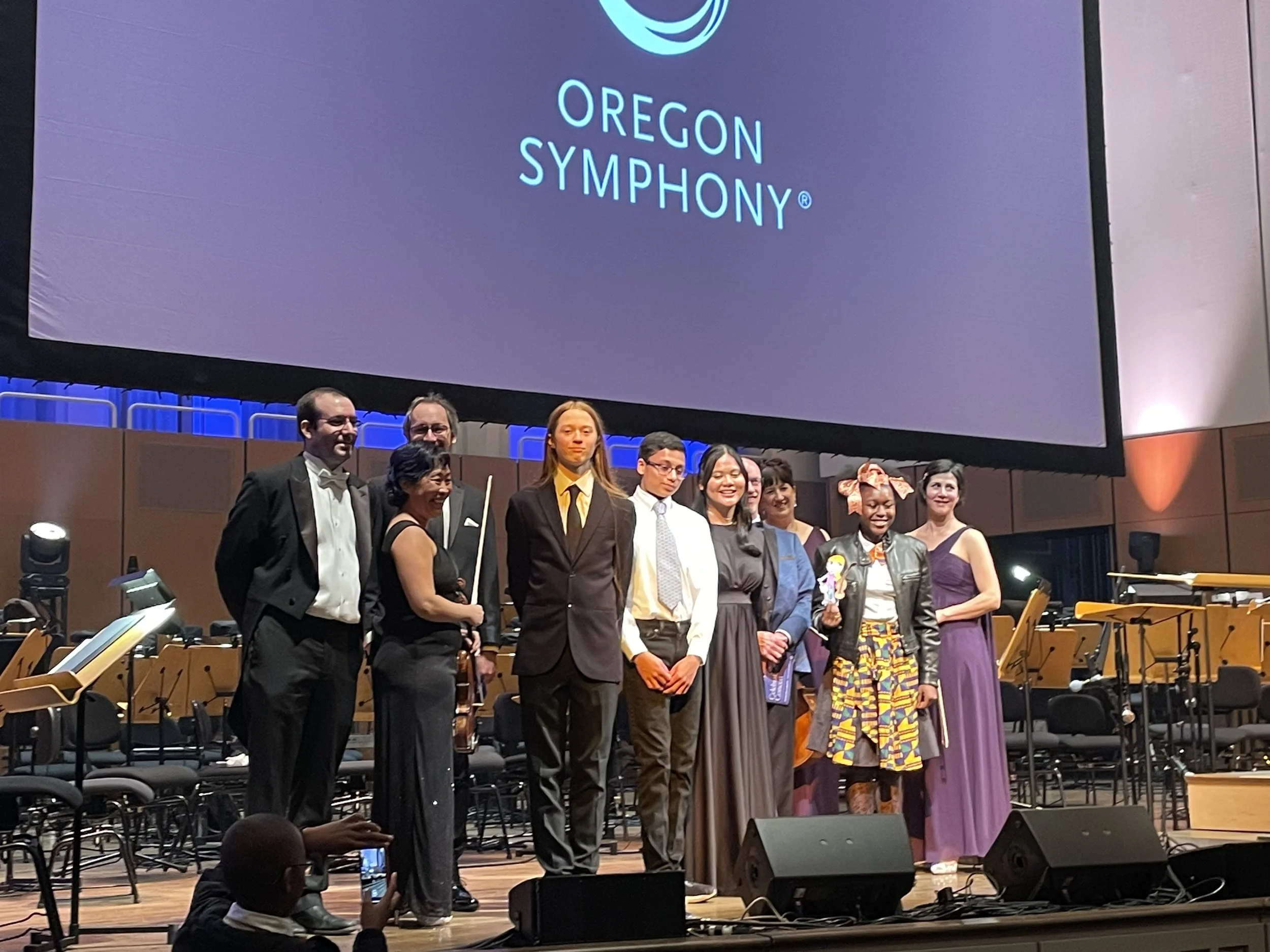 A group of nine people standing on stage after a performance, in front of a large screen with the Oregon Symphony logo.