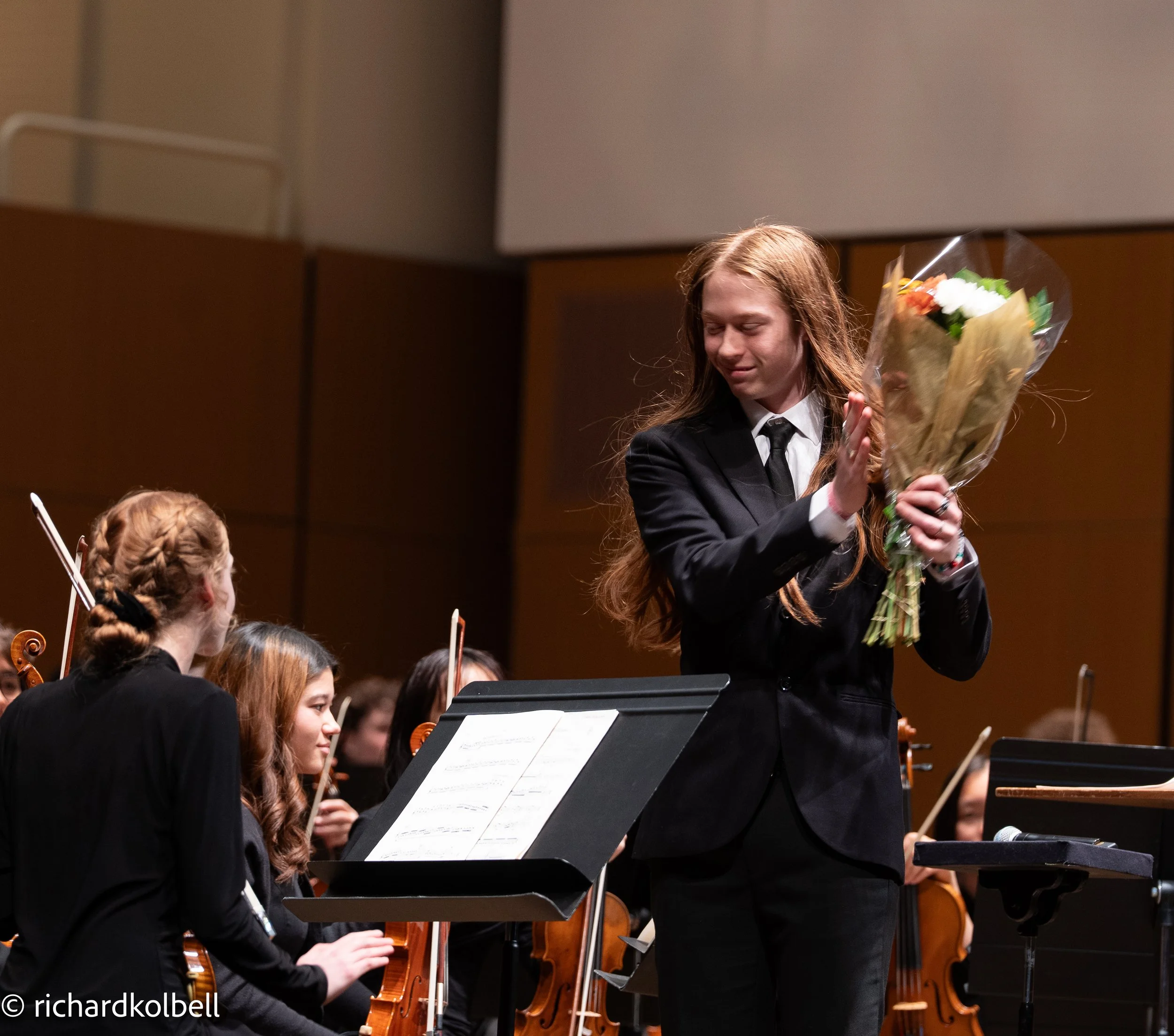 A young woman in a black suit and tie holding a bouquet of flowers on stage, while a group of young women with violins are seated behind her, in a concert hall.