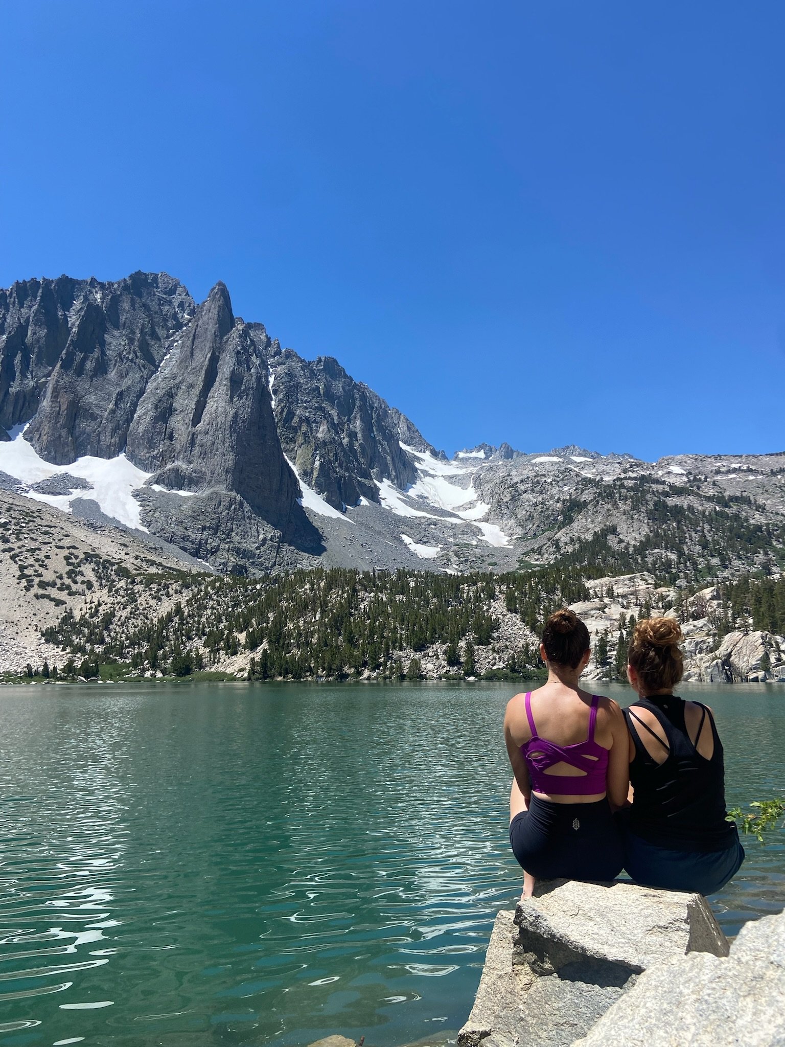Two women sitting on a rock by a lake with a mountain range, snow patches, and a clear blue sky in the background.