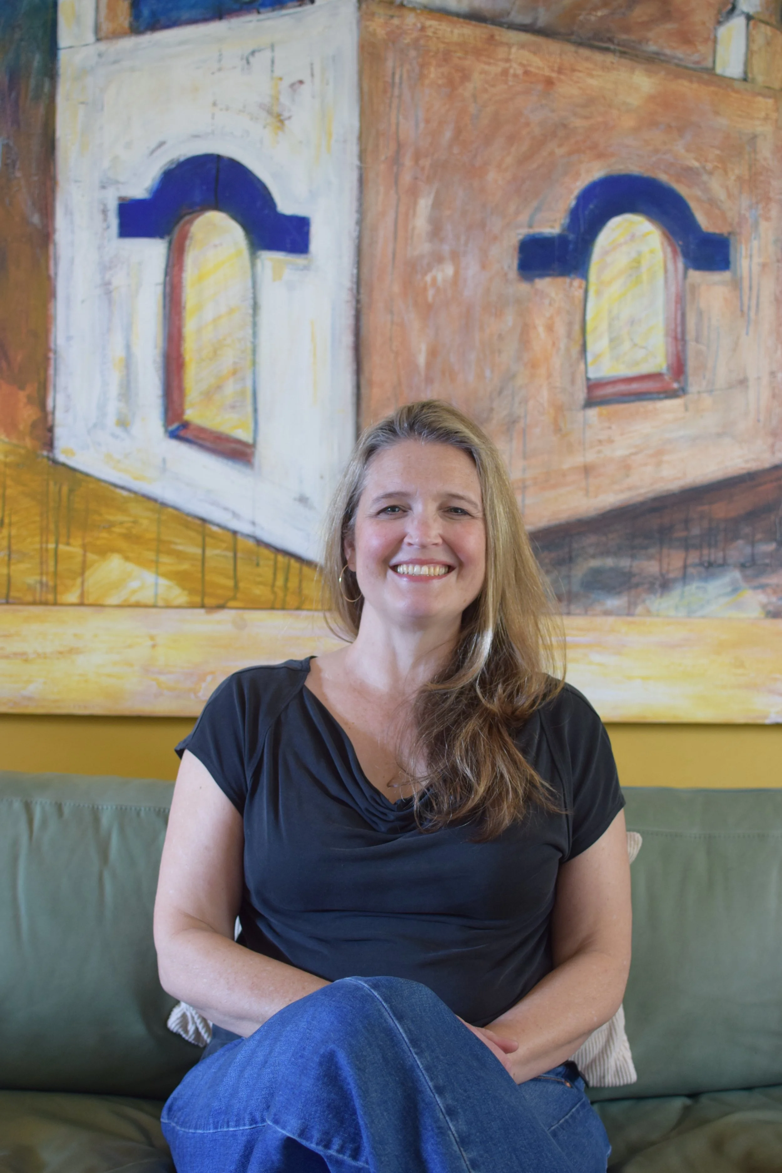 A woman with long, wavy brown hair sitting on a green sofa, smiling. Behind her is a large painting of a building with arched windows and shutters in warm colors.