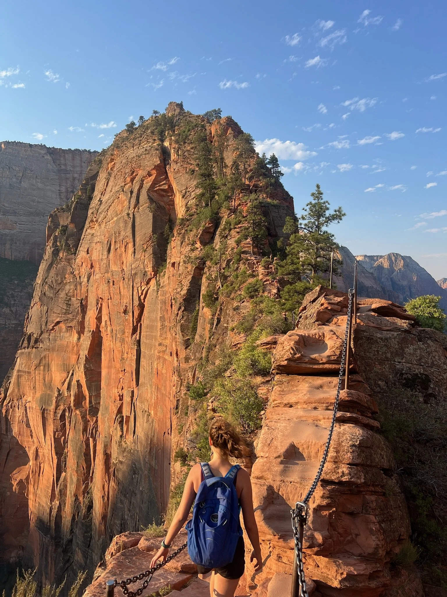 A woman with a blue backpack walking on a mountain trail with a chain fence, surrounded by red rock cliffs and green trees in a desert landscape under a blue sky.