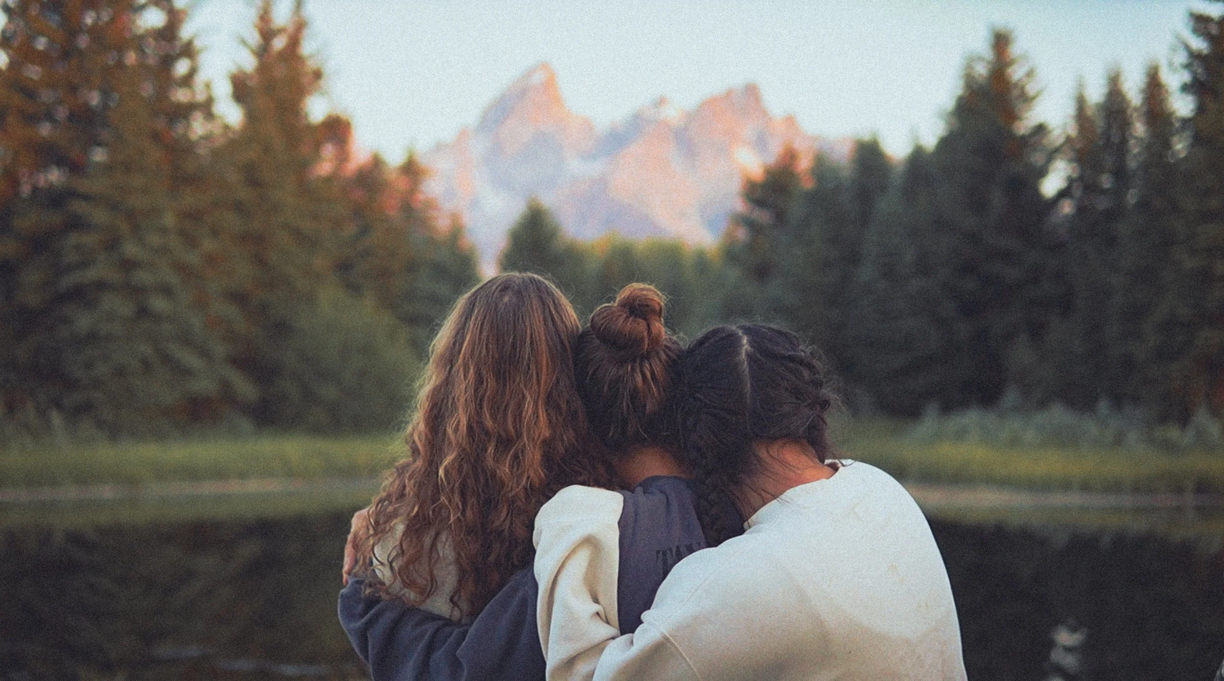 Three women sitting closely together outdoors, with their backs to the camera, overlooking a lake and mountains in the distance during sunset or sunrise.