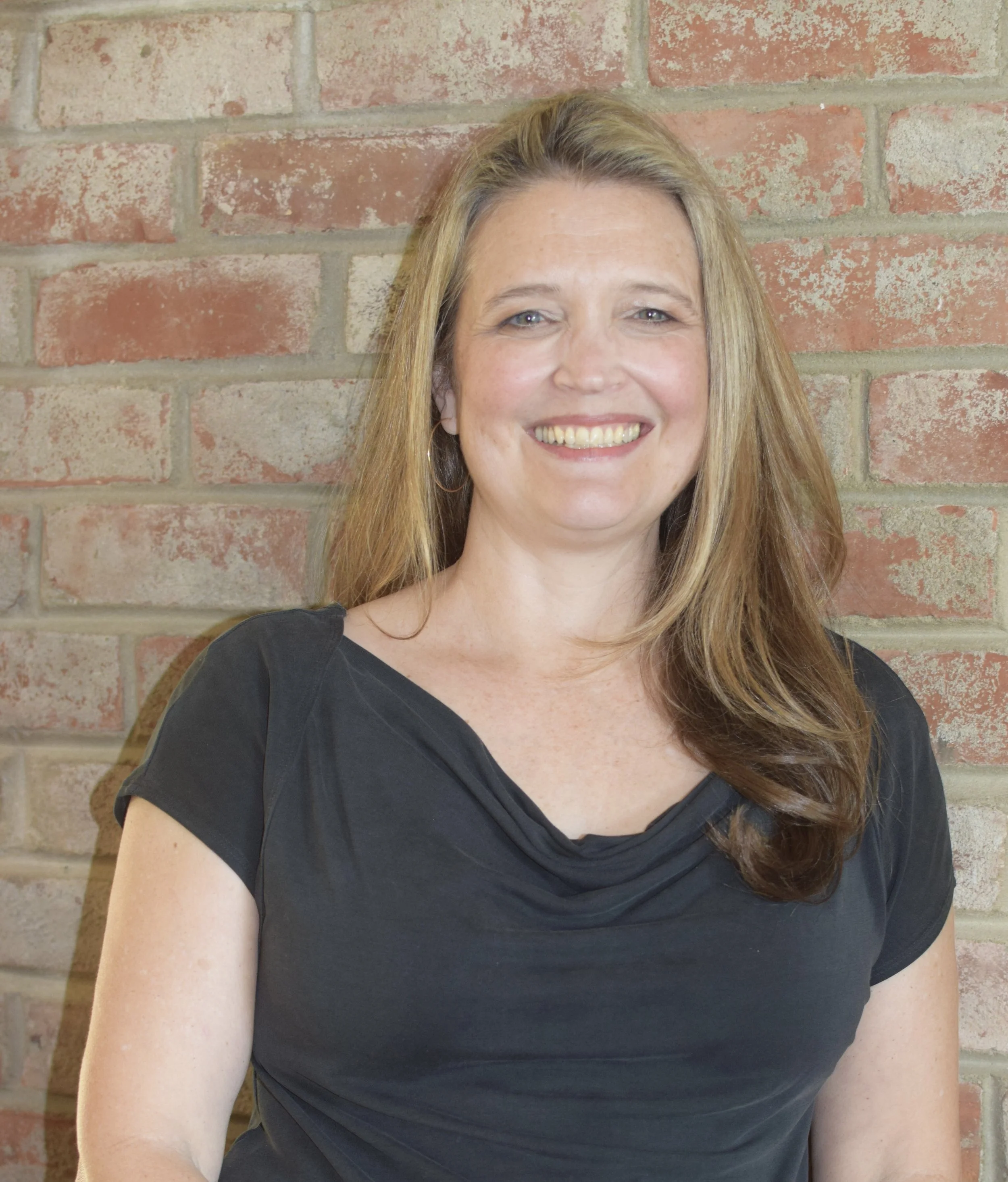 A woman with long, wavy, reddish-brown hair smiling in front of a brick wall.