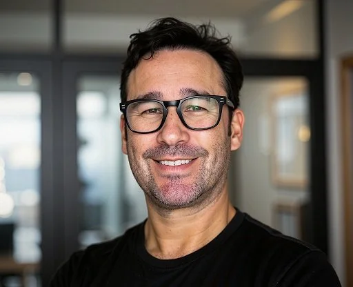 A smiling man with dark hair, glasses, and light facial hair, wearing a black shirt, standing indoors with windows and blurred background.