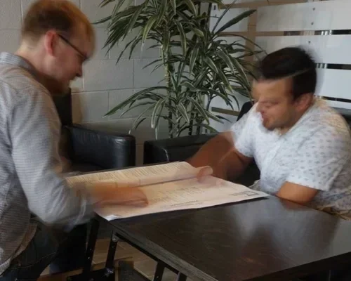 Two men sitting at a table in a cafe or restaurant, looking at a menu. One man has glasses and light-colored hair, wearing a checkered shirt. The other has dark hair and is wearing a white shirt. There is a large plant in the background.