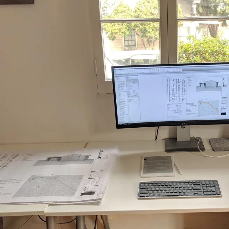 Desk with architectural blueprints, a computer monitor displaying architectural plans, a wireless keyboard, an e-reader, and a window showing greenery and a parked vehicle outside.