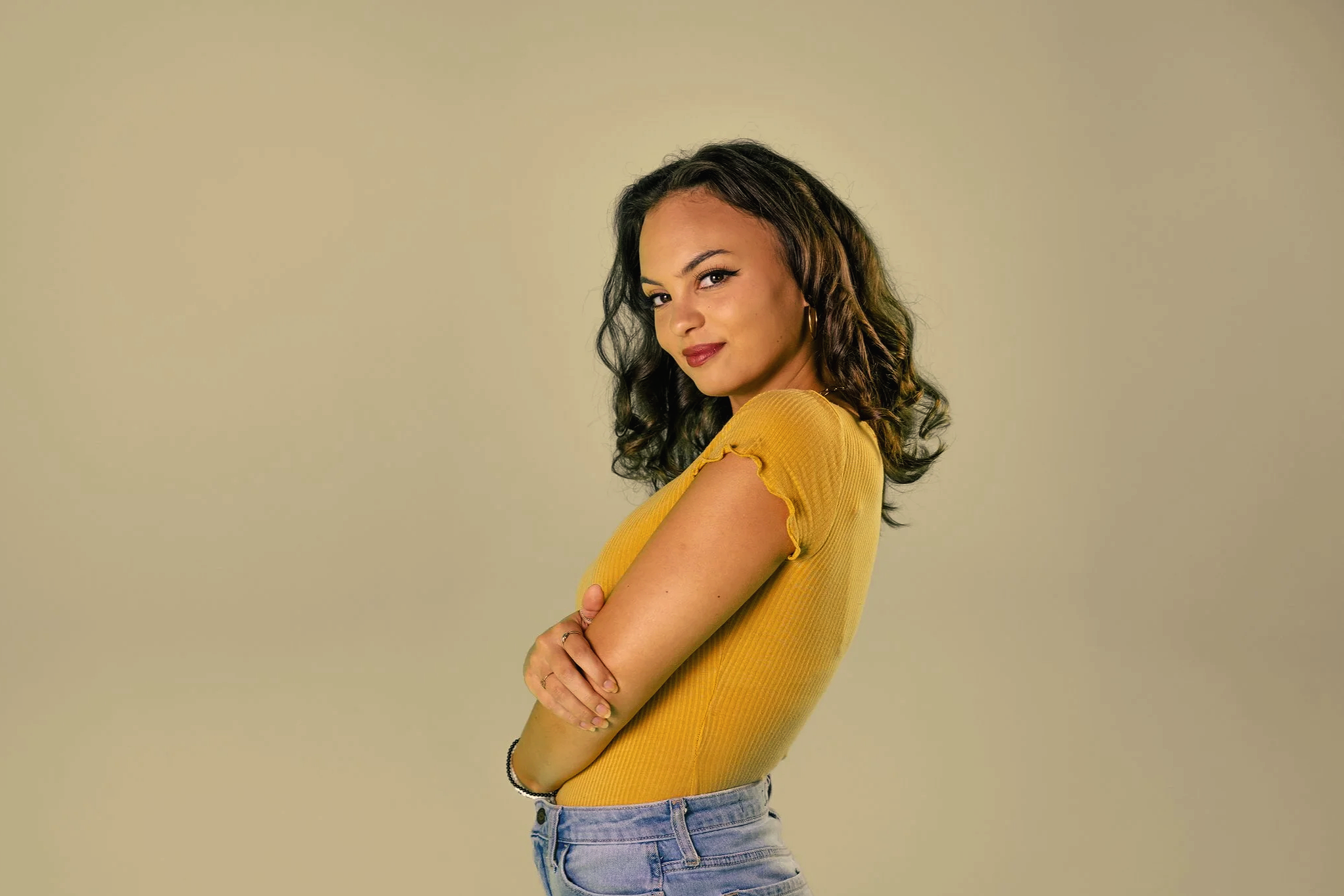 A young woman with dark, curly hair, wearing a yellow top and light blue jeans, standing against a plain, light-colored background. She has a confident expression, with arms crossed and a slight smile.