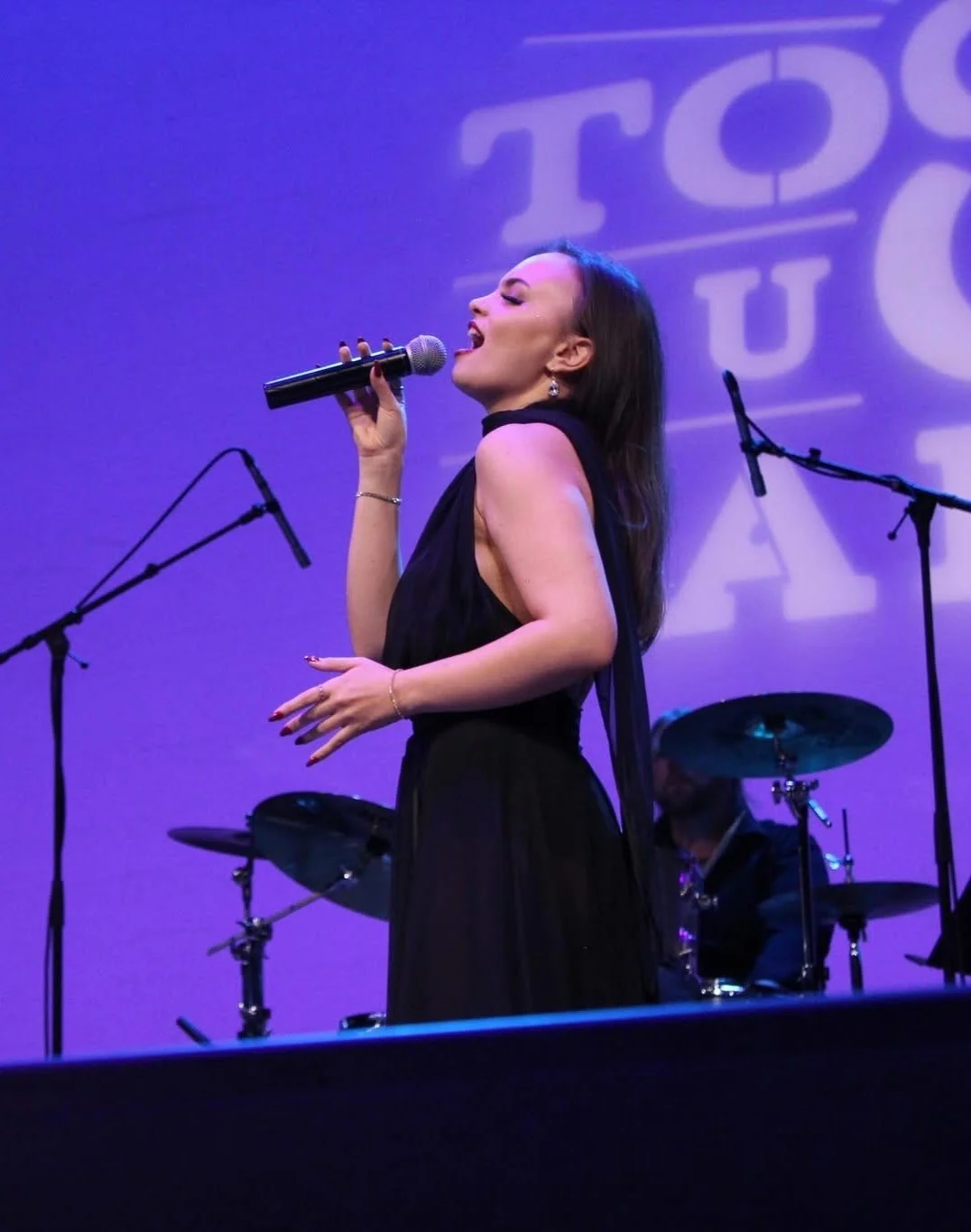 A woman singing on stage with a microphone in her hand, wearing a black dress, with purple lighting and a drum set in the background.