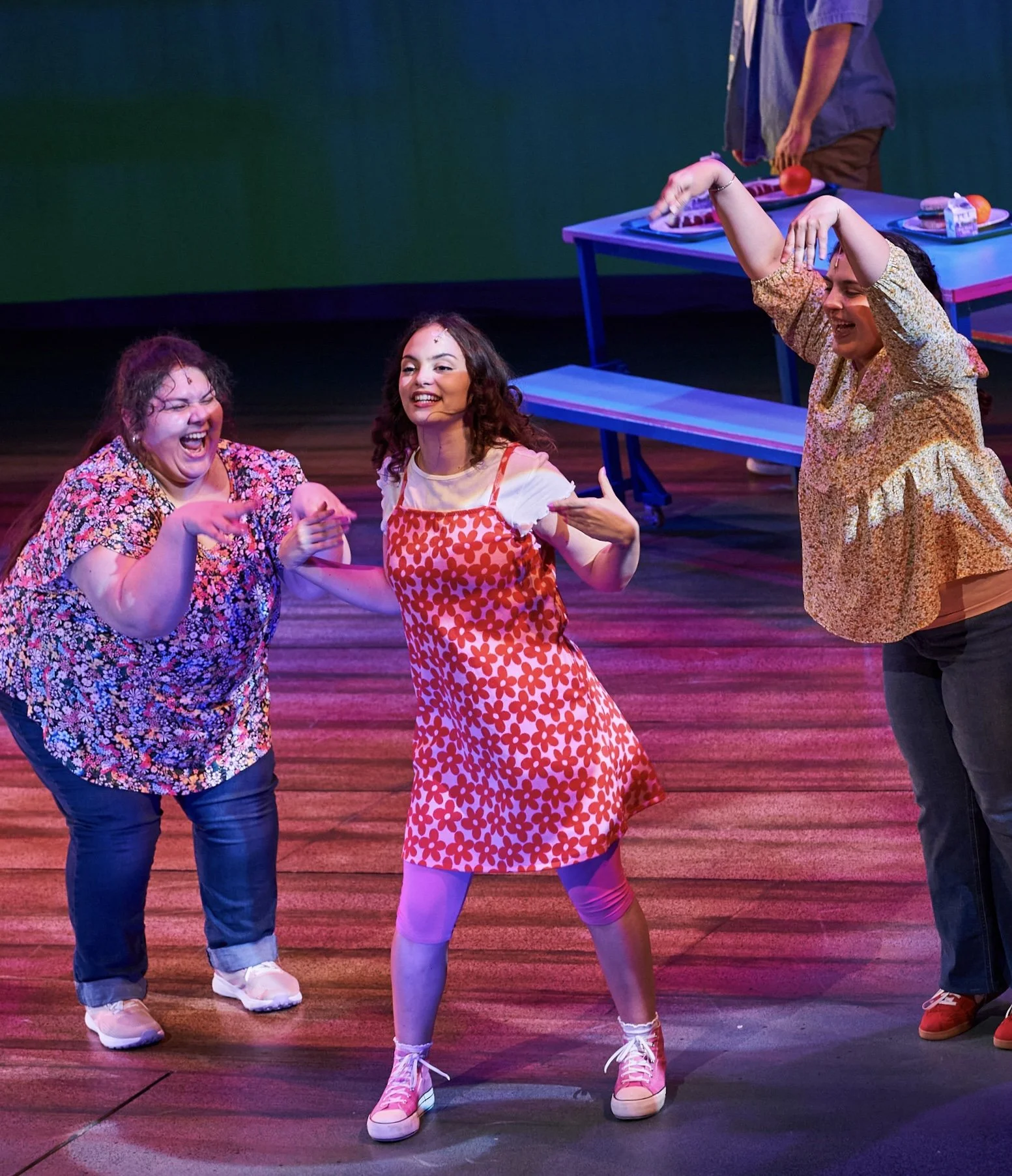 Three women laughing and dancing on stage during a theatrical performance, with a person behind a table with props in the background.
