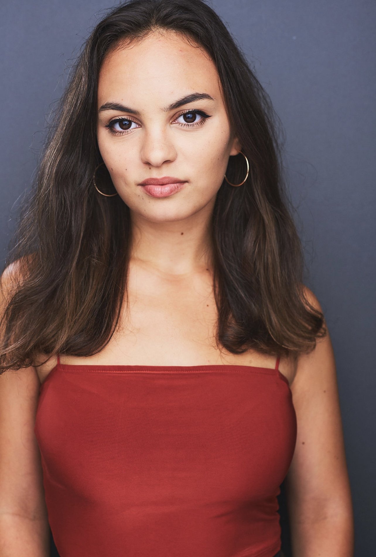 A young woman with long dark hair wearing a red spaghetti strap top and hoop earrings, standing against a dark gray background.