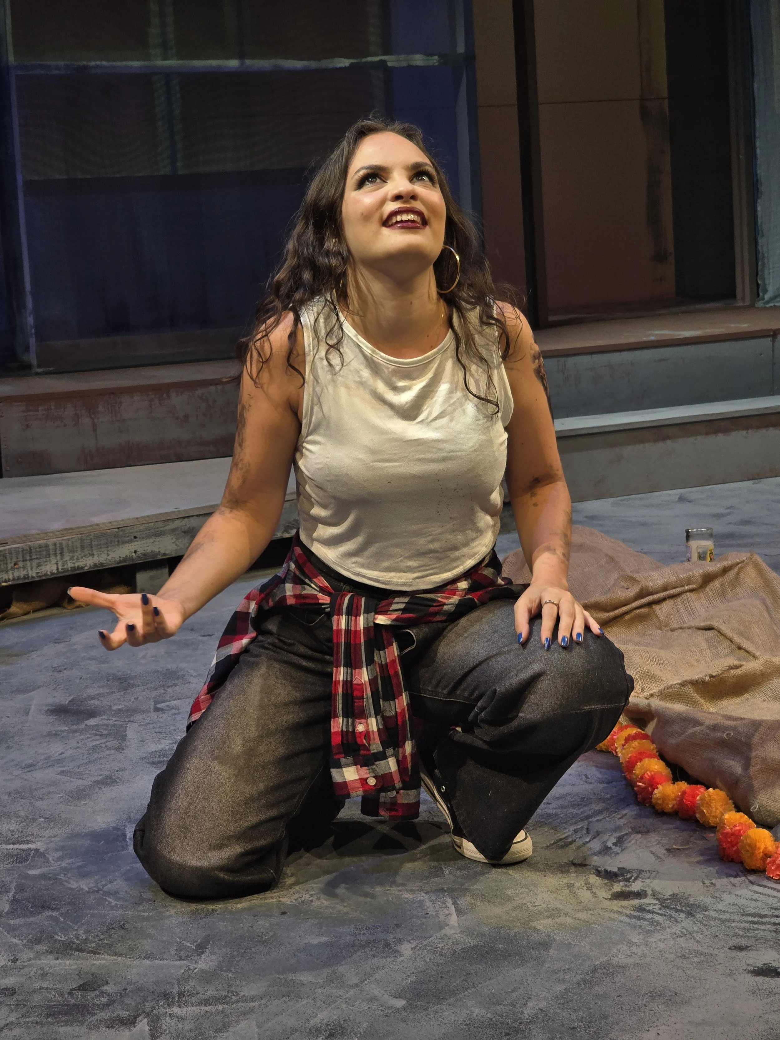 A woman kneeling on the floor, looking upwards with an expressive face. She has wavy dark hair, large hoop earrings, and is wearing a sleeveless beige top and black jeans with a red plaid shirt tied around her waist. There are colorful decorations an
