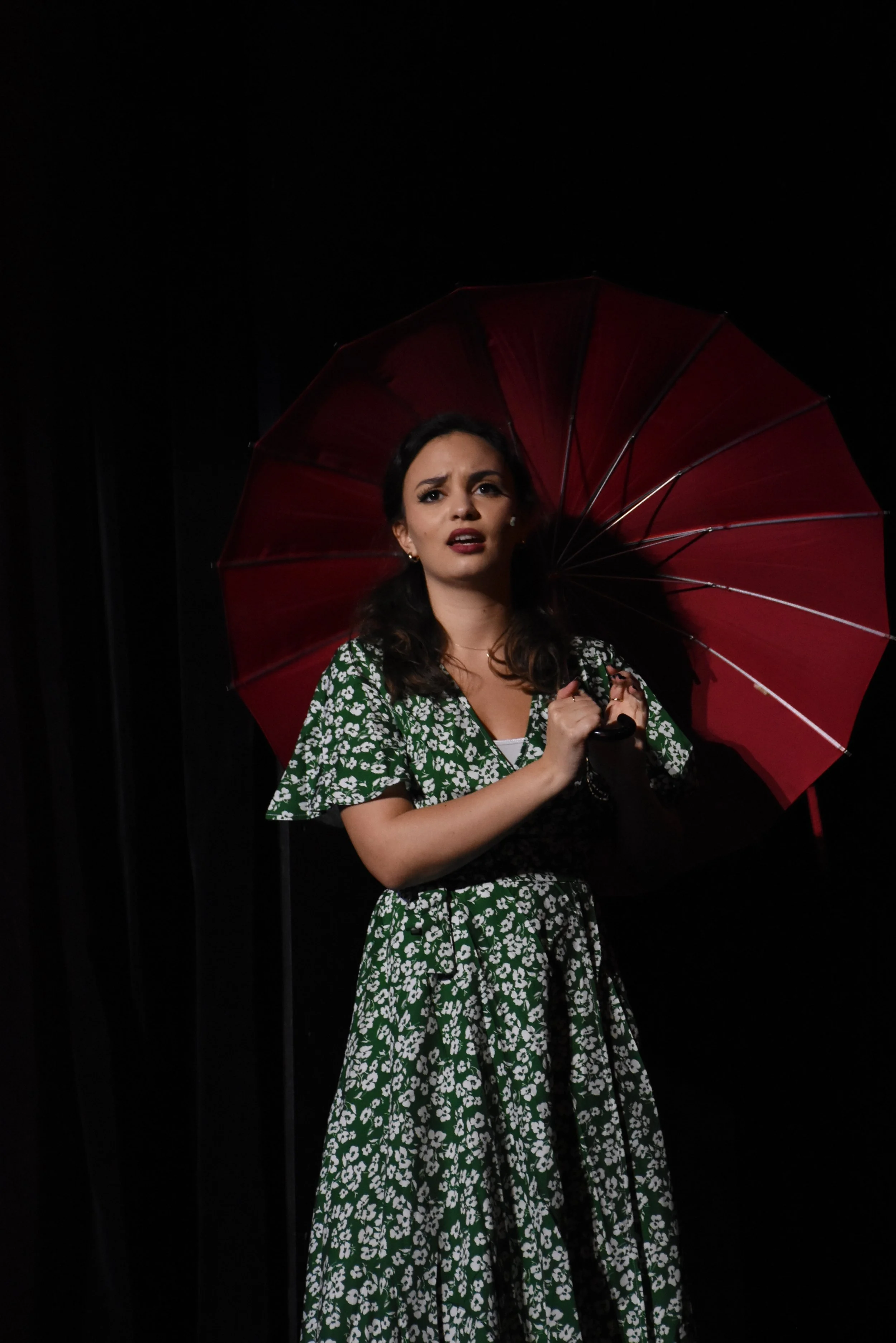 A woman with dark hair, wearing a green floral dress, holding a red umbrella, standing against a dark background.