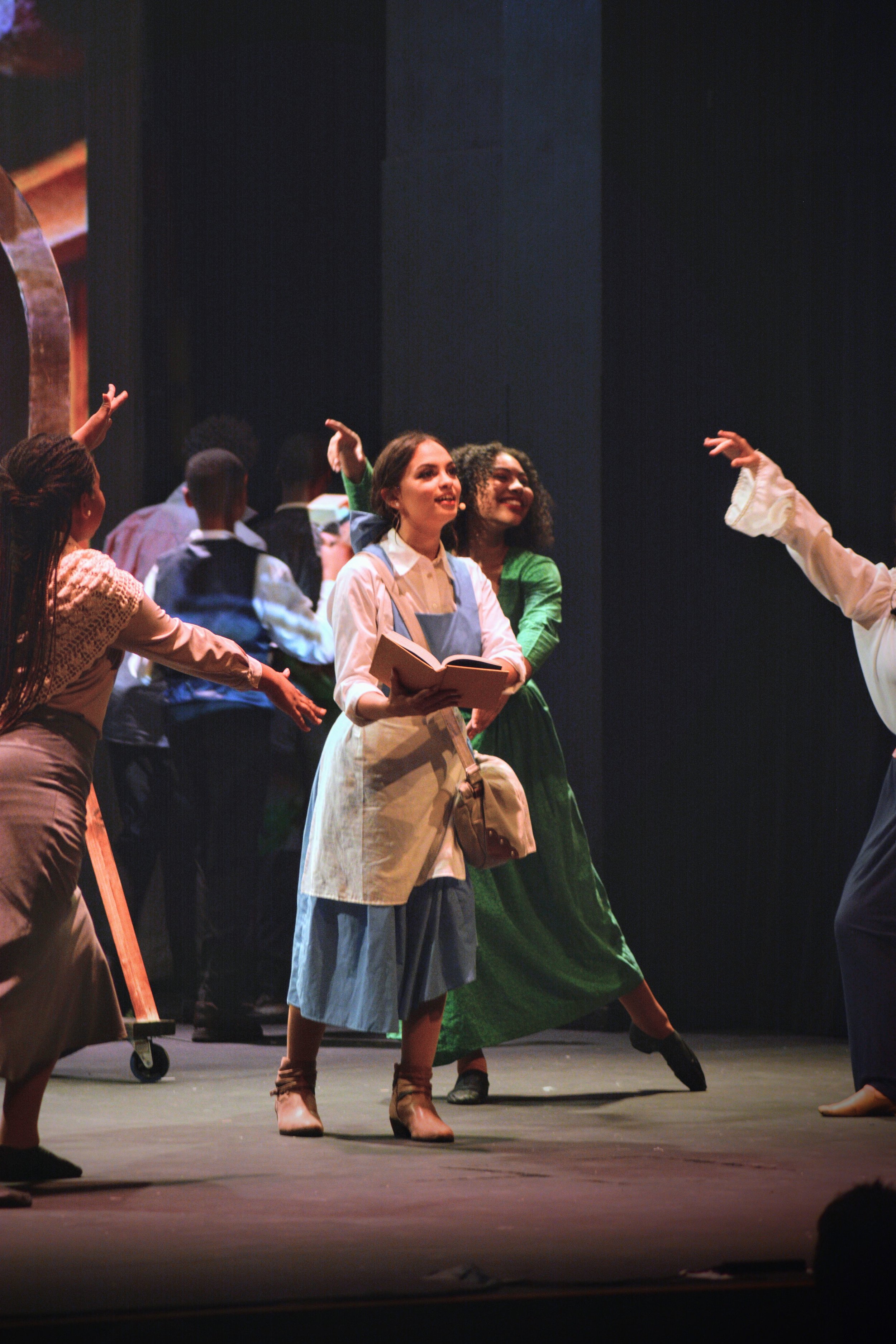 Actors performing on stage in a theatrical play or musical, dressed in period costumes, with some holding books or props.