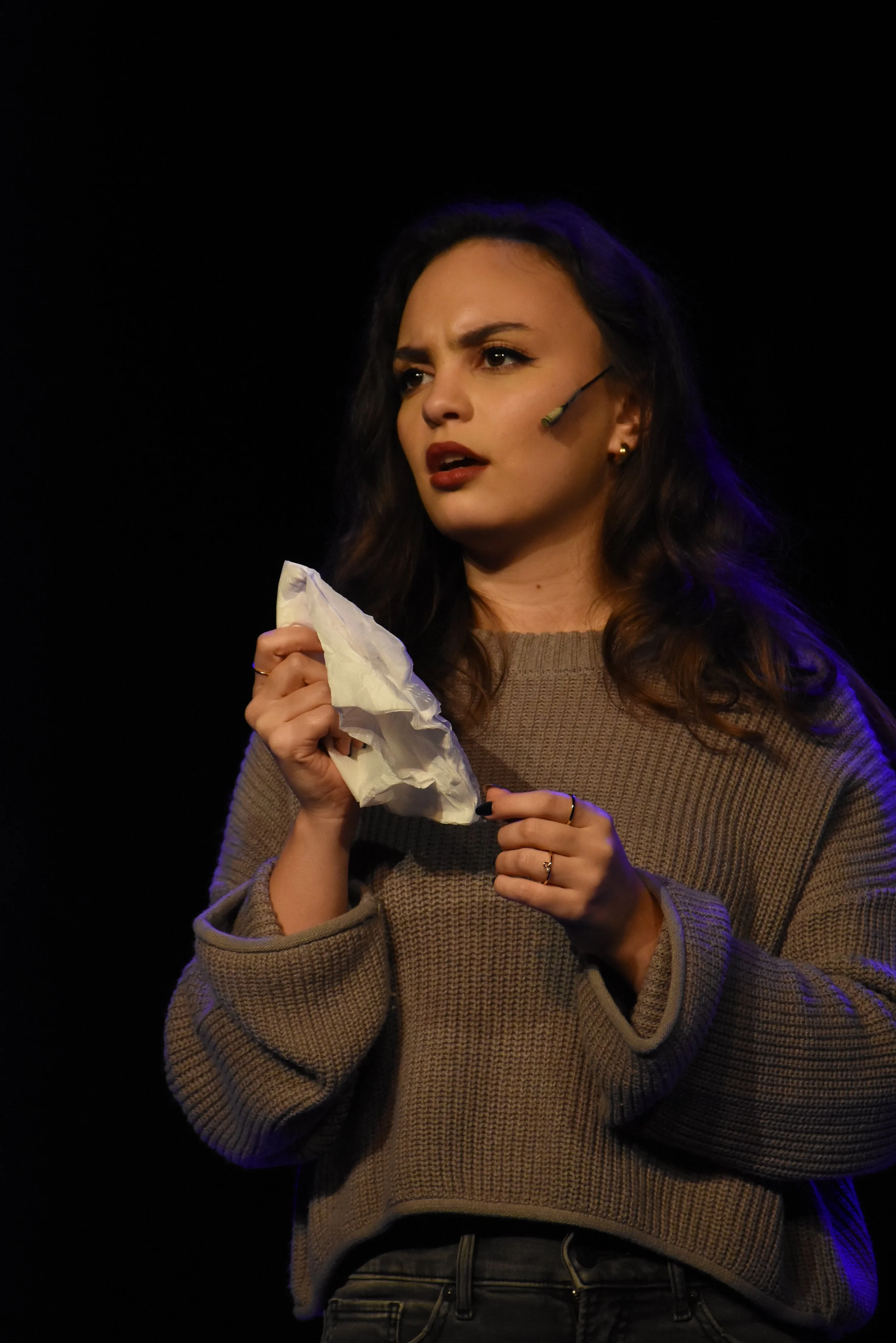 A young woman with dark wavy hair and light skin stands on a dark stage, holding a crumpled tissue in her right hand, with a thoughtful or concerned expression. She is wearing a beige sweater and has a headset microphone.