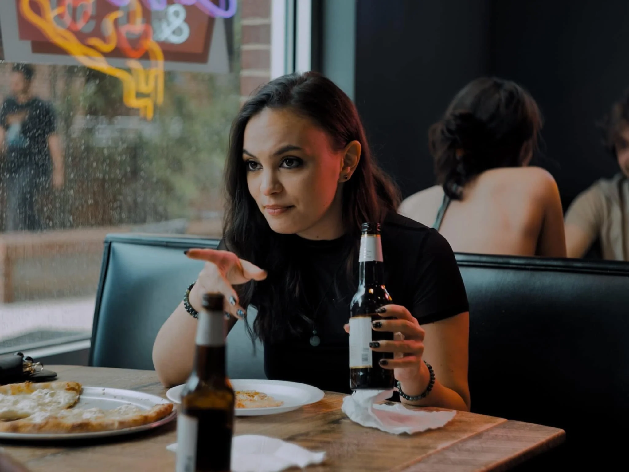 A woman with dark hair and a black shirt sitting at a table in a restaurant, holding a beer bottle and gesturing with her hand, with pizza on a plate in front of her, and other people sitting in the background.