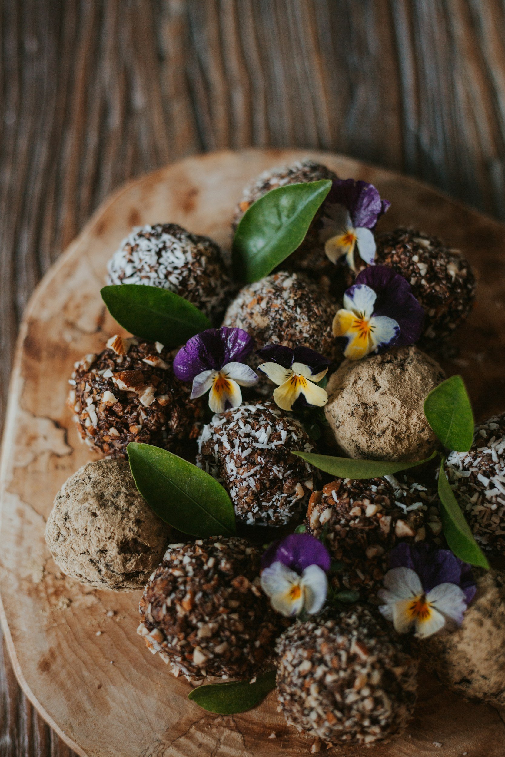 Assorted coconut-covered chocolate truffles on a wooden platter, decorated with purple and yellow edible flowers and green leaves.