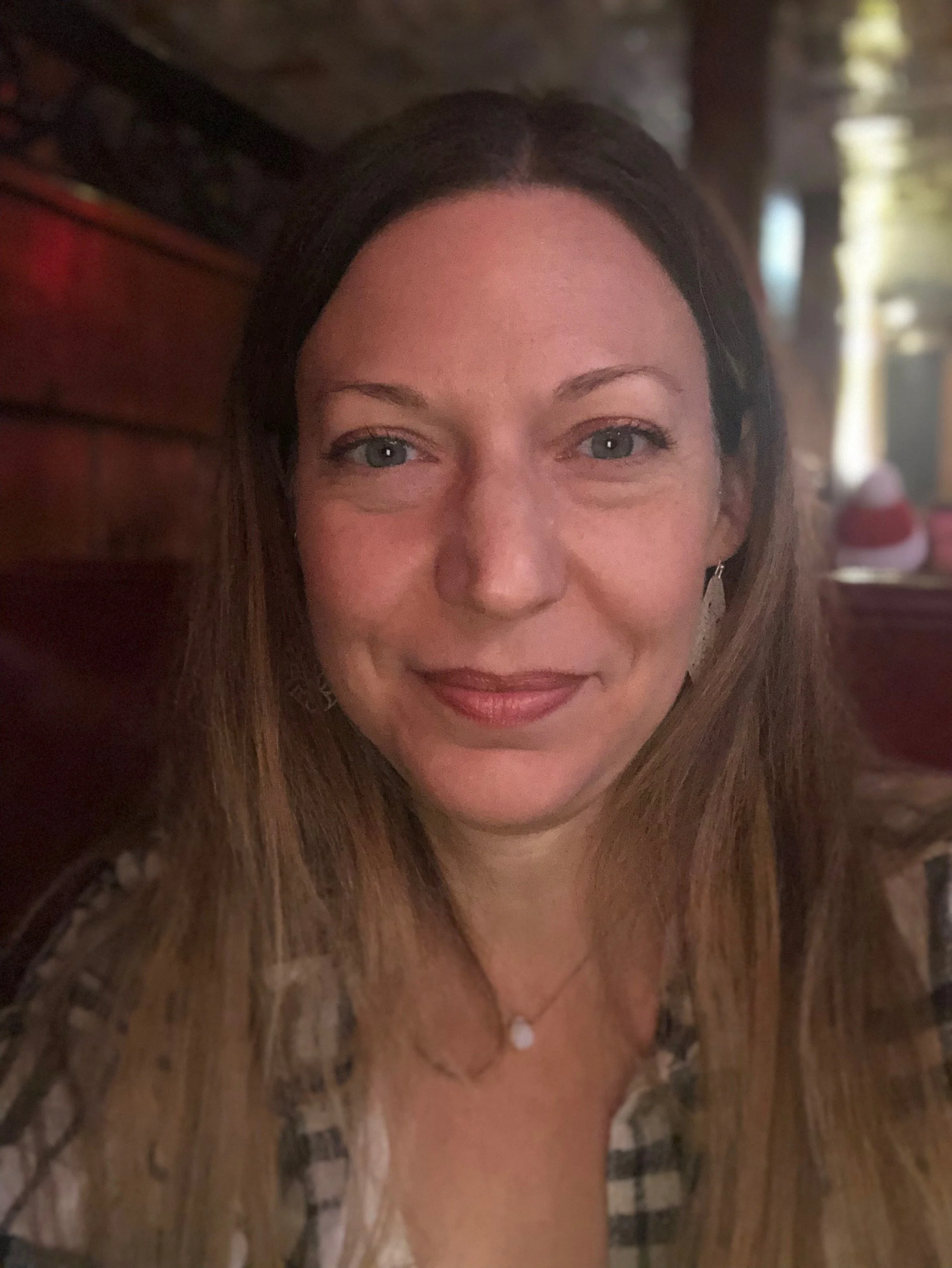 A smiling woman with long brown hair and blue eyes, wearing earrings and a necklace, sitting in a restaurant with warm lighting and wooden decor.