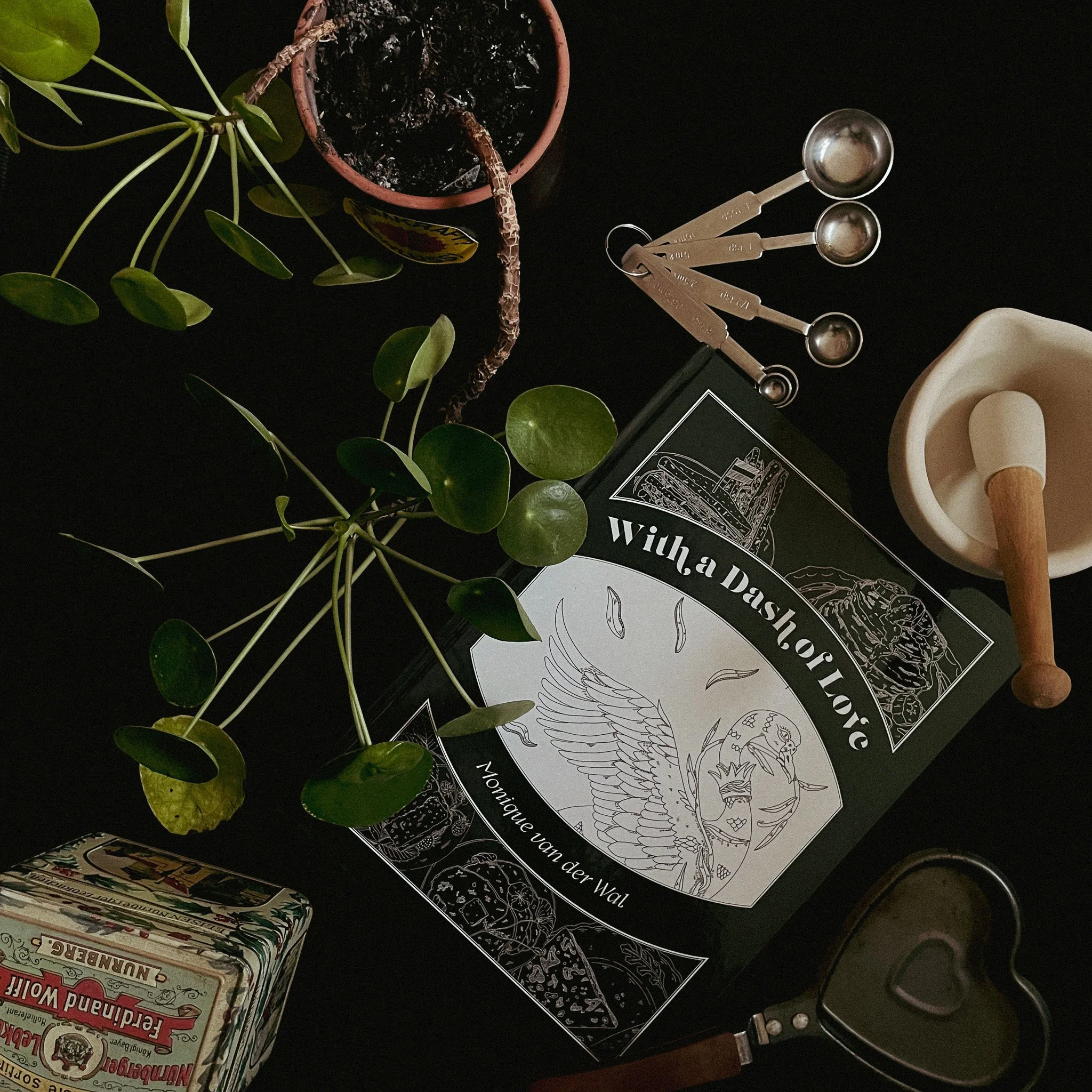 Top-down view of a collection of objects on a black surface, including a plant with round green leaves, a small open tin of soil, a book titled 'With a Dash of Love' by Monique Van der Wal, a set of metal measuring spoons, a white mortar and pestle, an illustrated tin box, and a metal heart-shaped mold.