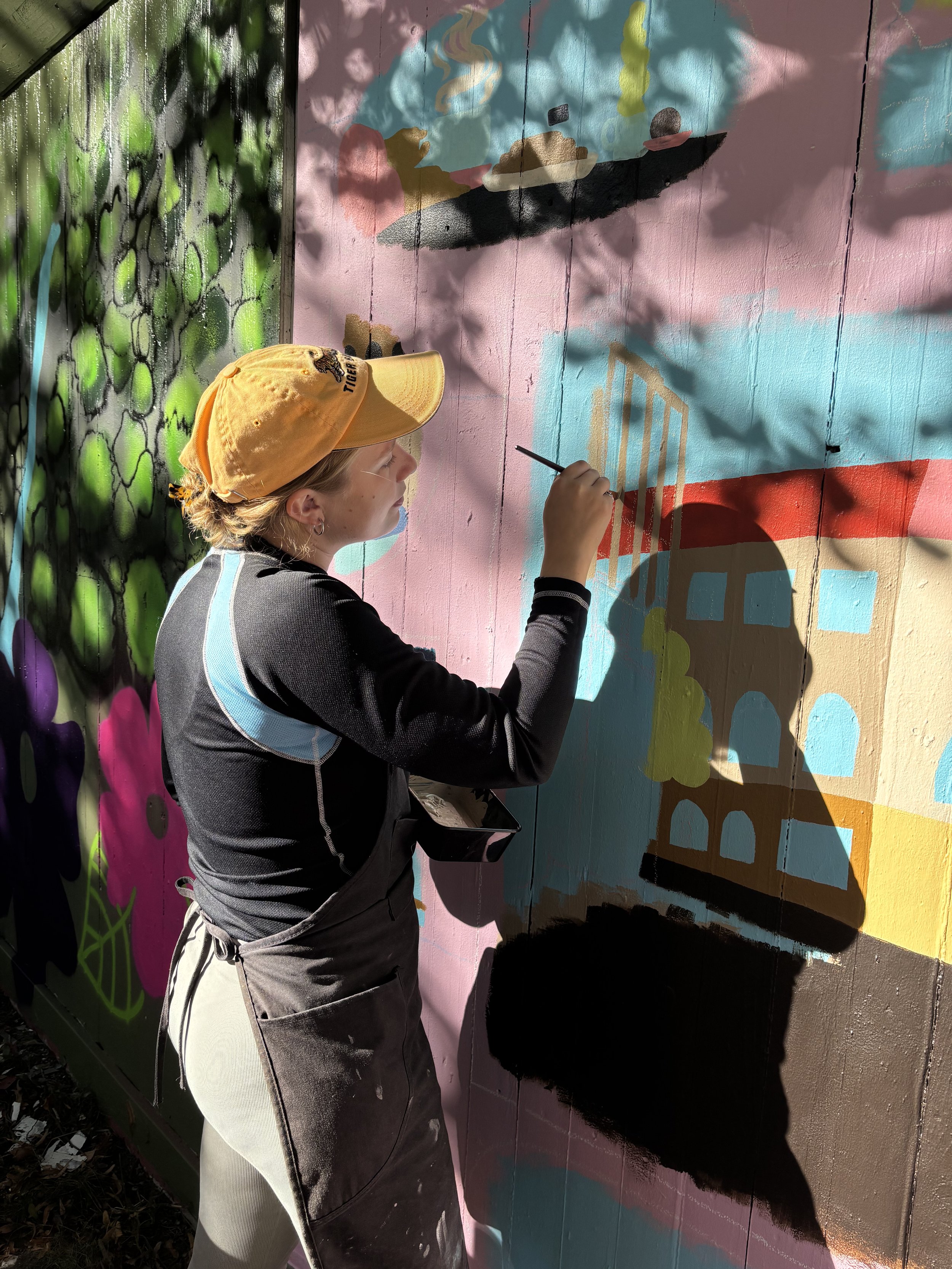 A girl wearing a yellow baseball cap and aprons painting a colorful mural on a wall, featuring a building, clouds, and abstract shapes.